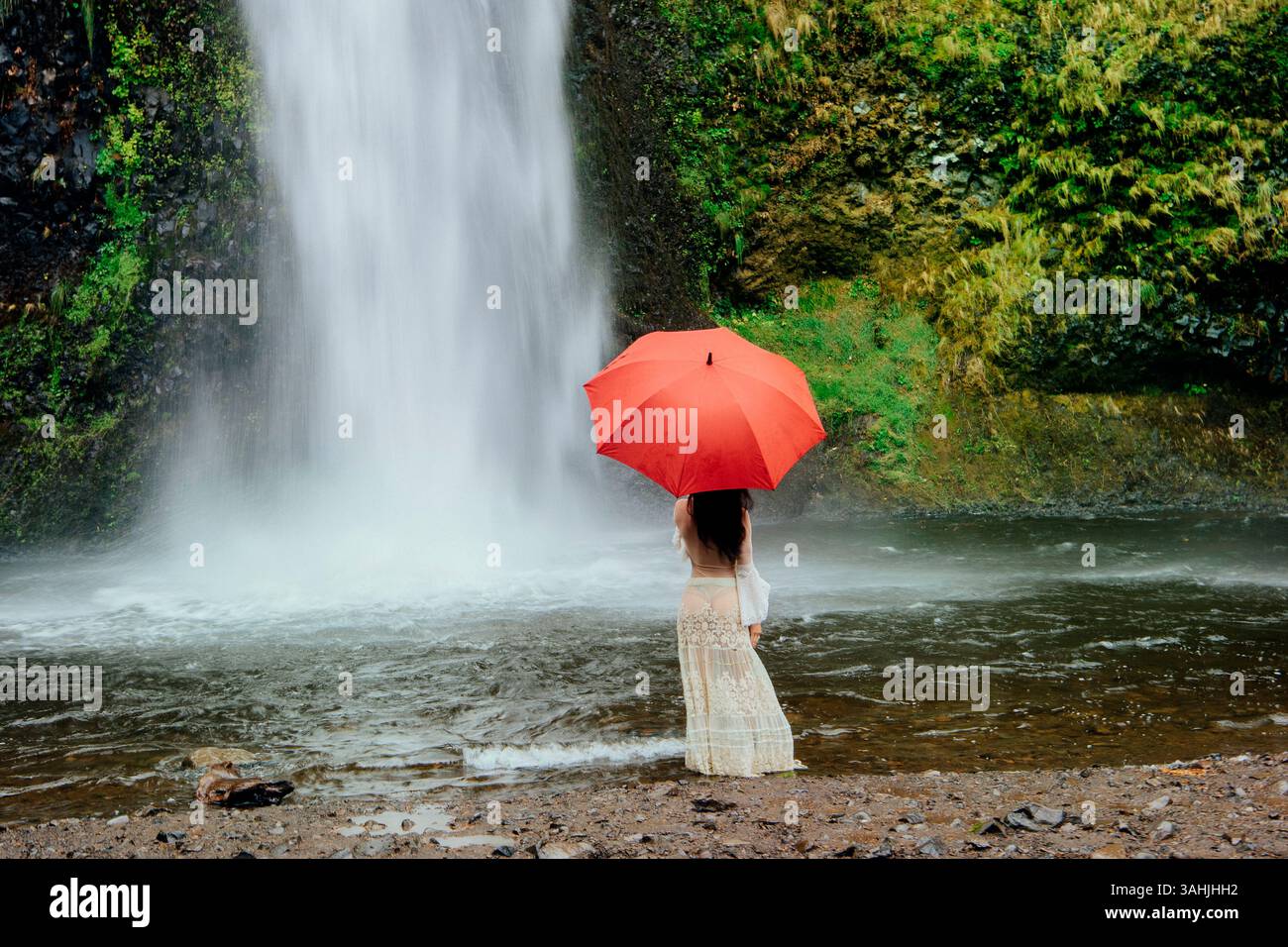 Donna con ombrello rosso si trova vicino a una cascata circondata da una vegetazione lussureggiante. Silver Falls, Oregon, Stati Uniti Foto Stock