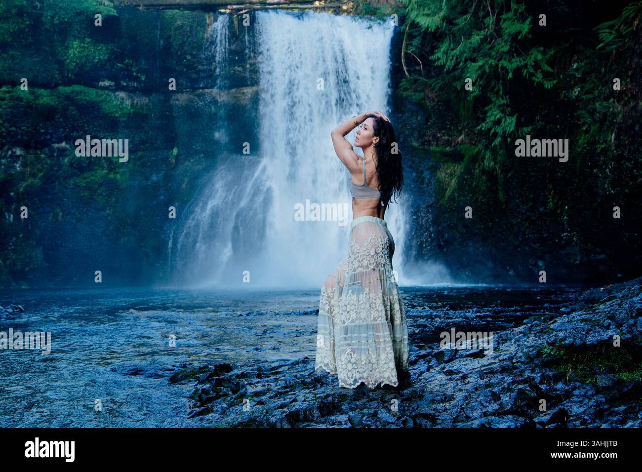 Una donna con una gonna bohémien si posa accanto a una maestosa cascata in una lussureggiante foresta verde. Silver Falls, Oregon, Stati Uniti Foto Stock
