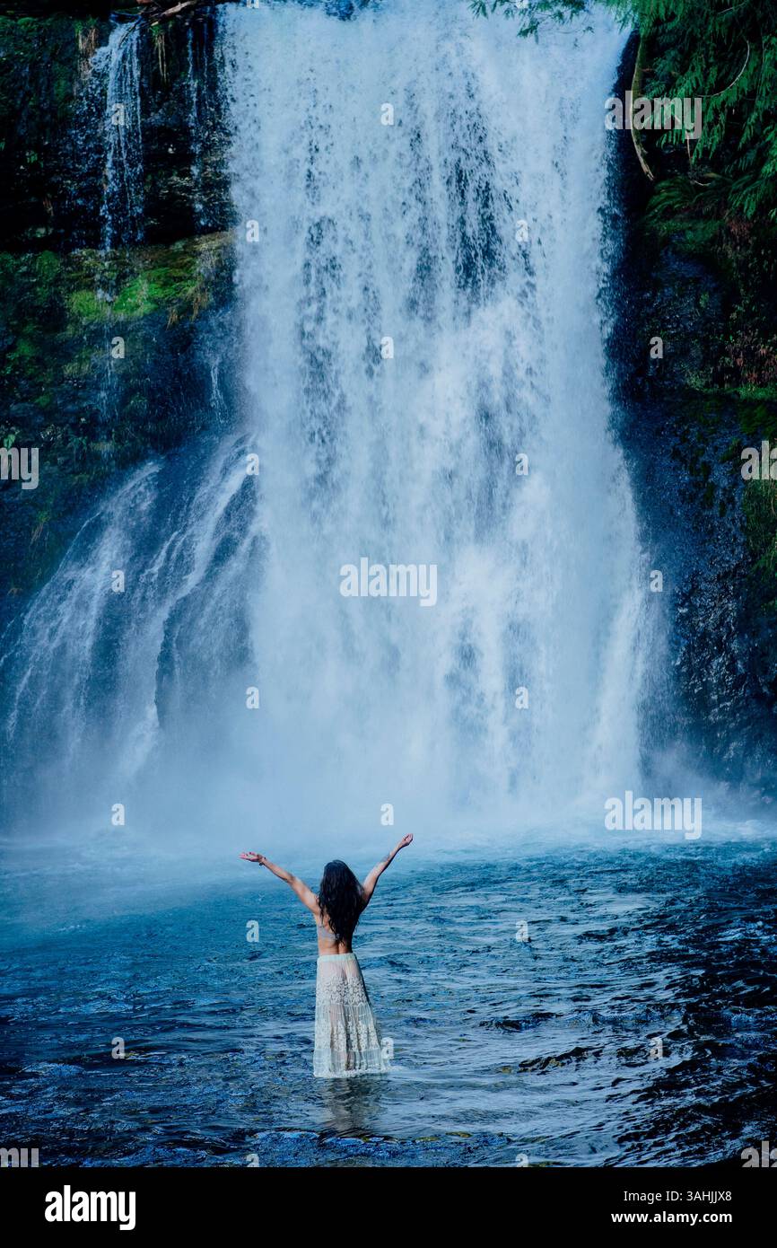 Donna in piedi in acqua con le braccia sollevate davanti a una grande cascata. Silver Falls, Oregon, Stati Uniti Foto Stock