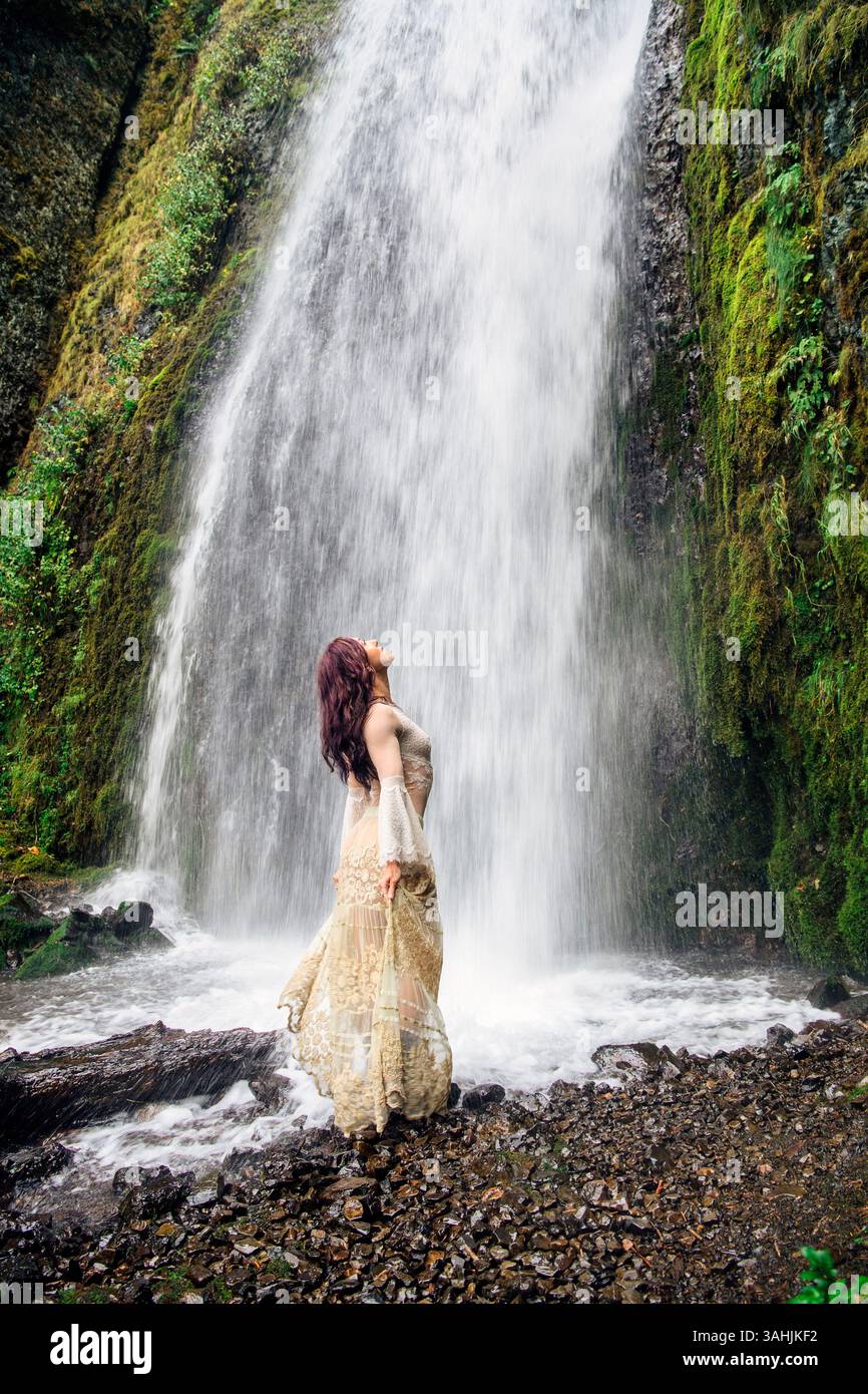Donna in un abito fluente in piedi alla base di una lussureggiante cascata verde. Silver Falls, Oregon, Stati Uniti Foto Stock