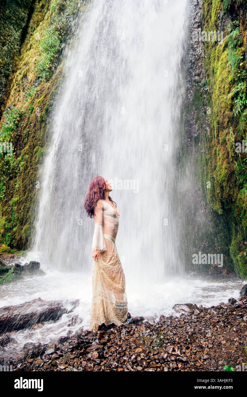 Donna in un abito fluente in piedi serenamente di fronte a una lussureggiante cascata. Silver Falls, Oregon, Stati Uniti Foto Stock