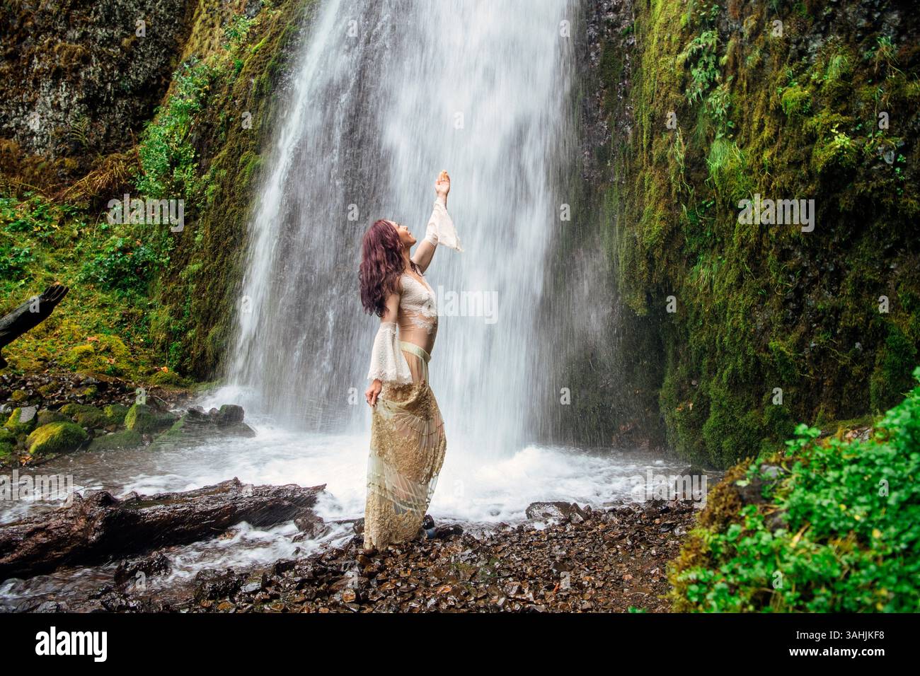 Una donna con un abito fluente posa graziosamente da una lussureggiante cascata circondata da una vibrante vegetazione. Silver Falls, Oregon, Stati Uniti Foto Stock