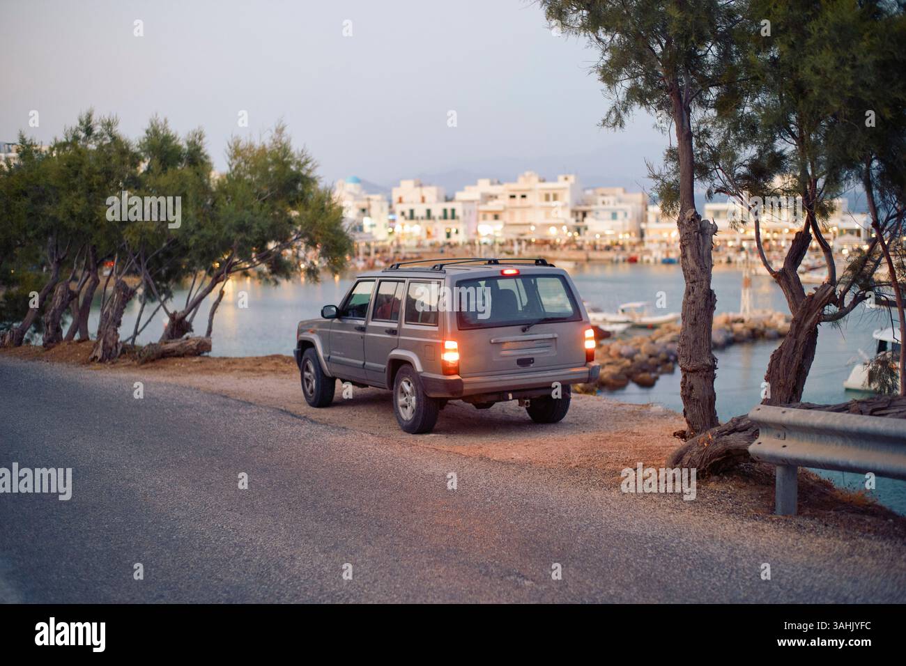 SUV parcheggiato da una strada costiera con edifici e barche sullo sfondo al crepuscolo. Mykonos, Grecia Foto Stock