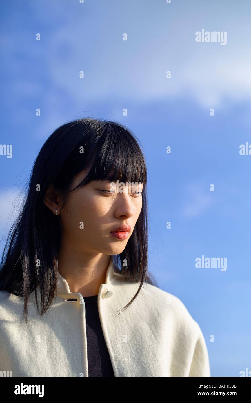 Donna con capelli neri e cappotto bianco si staglia contro un cielo blu con gli occhi leggermente chiusi. Brandeburgo, Germania Foto Stock
