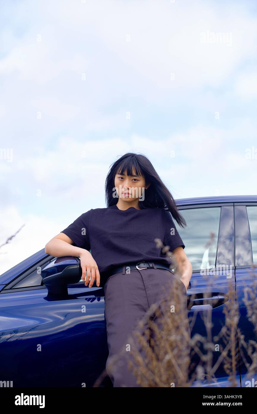 Donna in camicia nera si appoggia su un'auto blu scuro sullo sfondo di un cielo nuvoloso. Brandeburgo, Germania Foto Stock