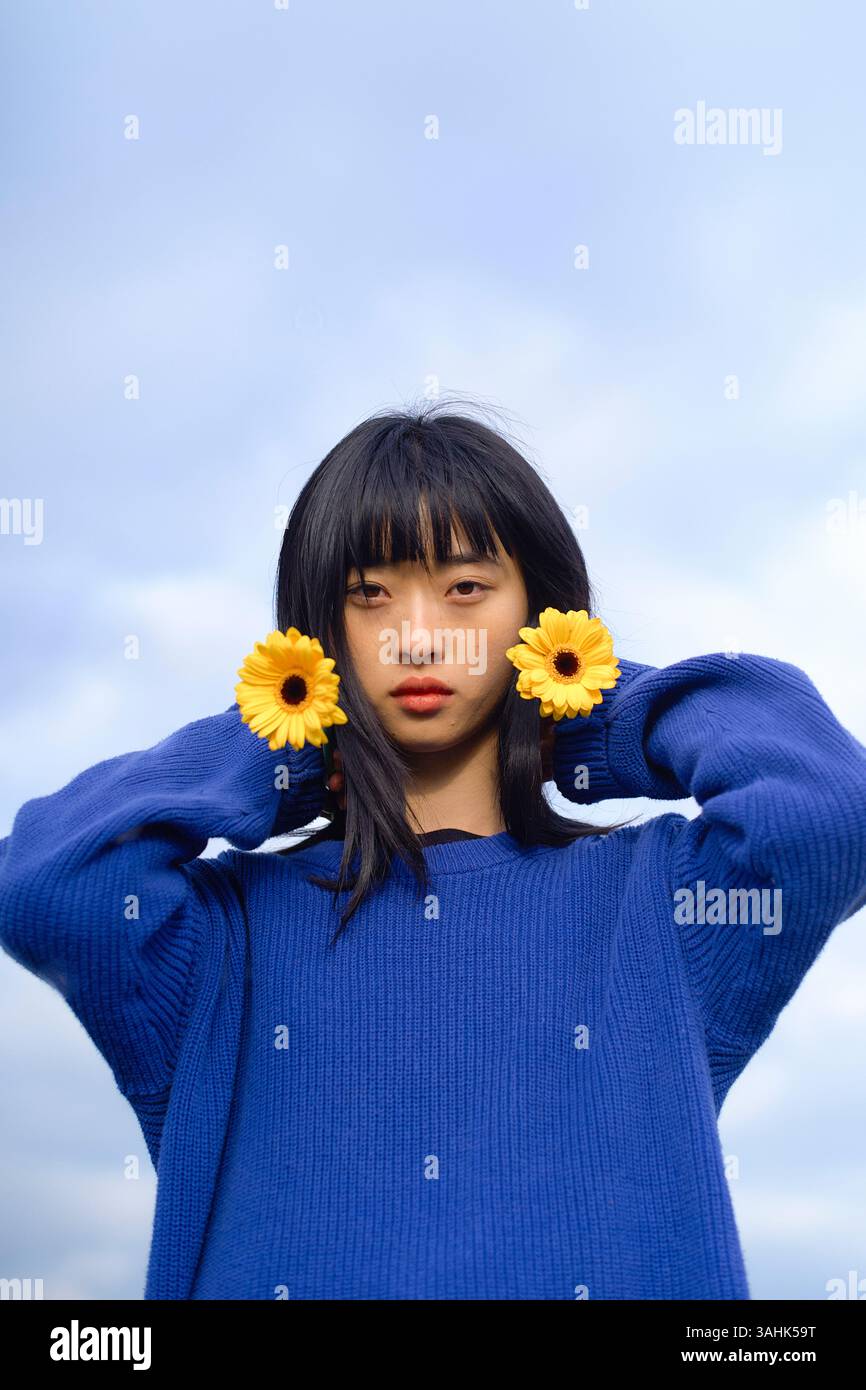Una donna con maglione blu tiene i fiori gialli alle orecchie contro un cielo nuvoloso. Brandeburgo, Germania Foto Stock