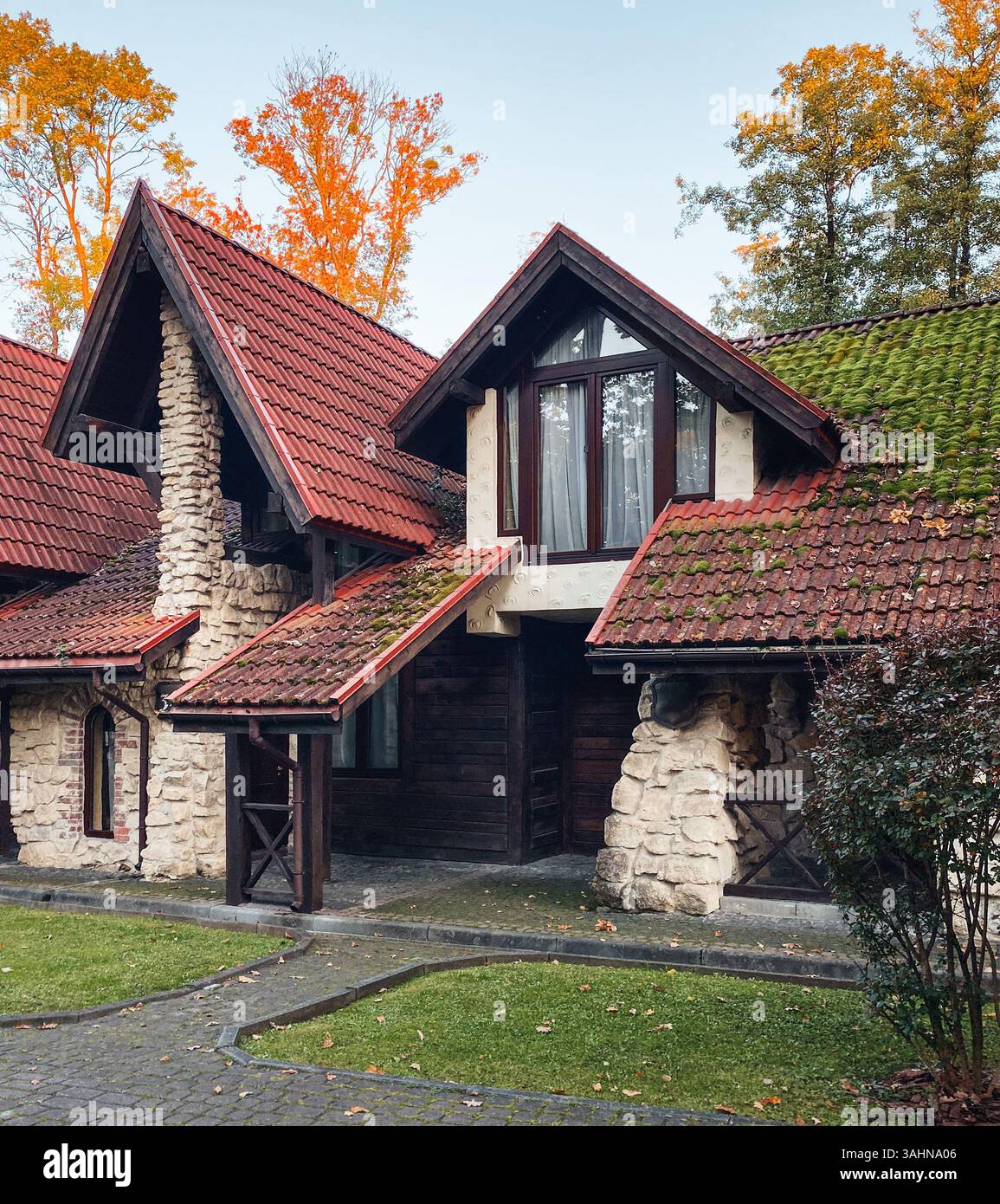 Due case rivestite di pietra in un boschetto di querce. Case a file con tetto in piastrelle rosse ricoperte di muschio circondate da alti alberi in un giorno d'autunno. Foto Stock