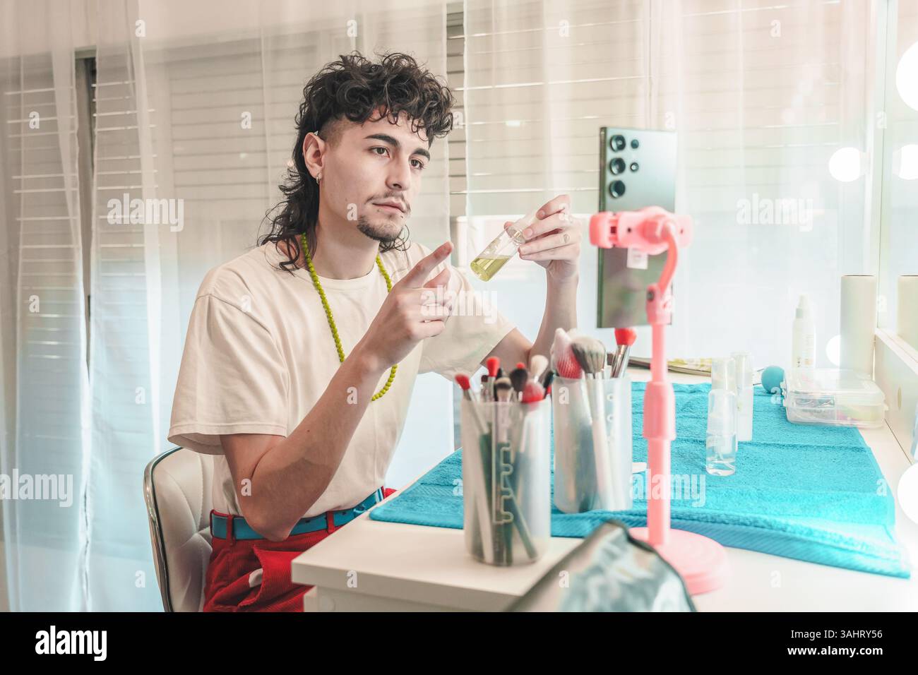 Giovane influencer di bellezza maschile latino che mostra il prodotto per la cura della pelle mentre registra video per i social media utilizzando lo smartphone a casa, promuovendo i cosmetici fino all'alto Foto Stock