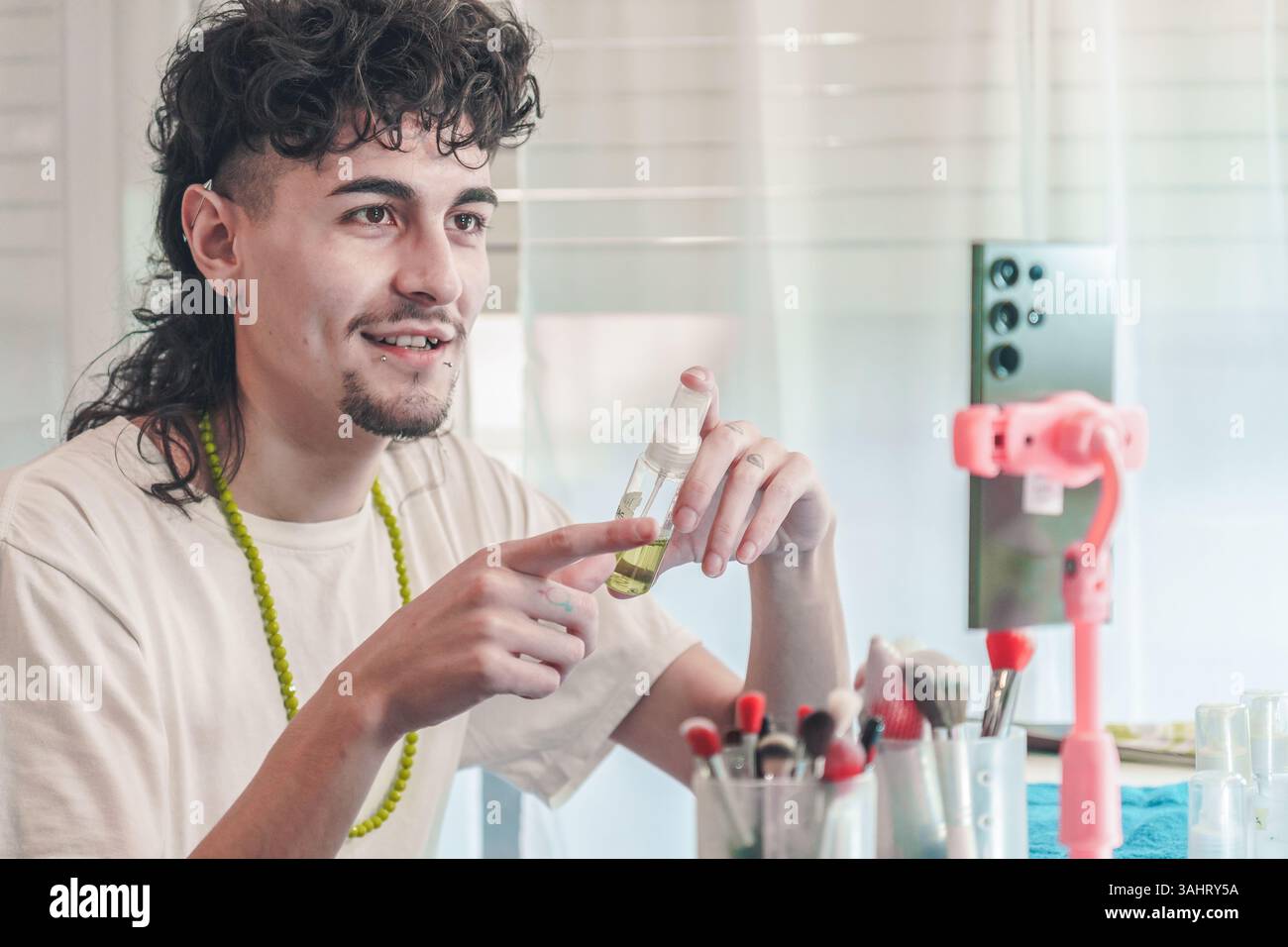 Giovane creatore di contenuti maschili che mostra una bottiglia di siero di bellezza durante la registrazione di video per i follower dei social media, promuovendo il marchio cosmetico e condividendo Make Foto Stock
