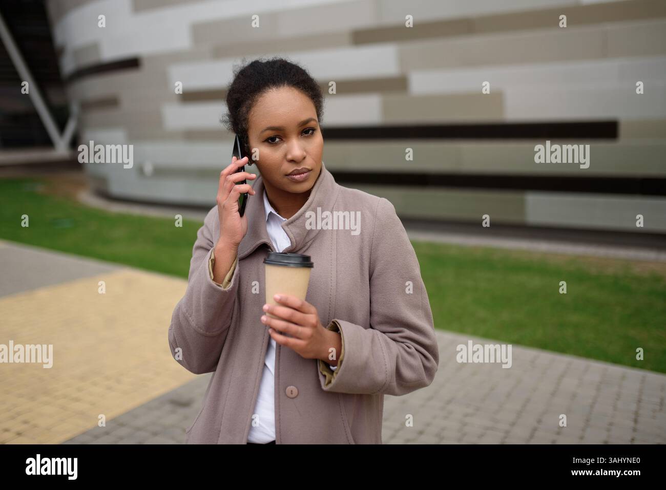Donna afroamericana seria in camice in piedi per strada in città, parlando al cellulare, tenendo una tazza di caffè in altra mano. Concetto di persone in città. Foto Stock