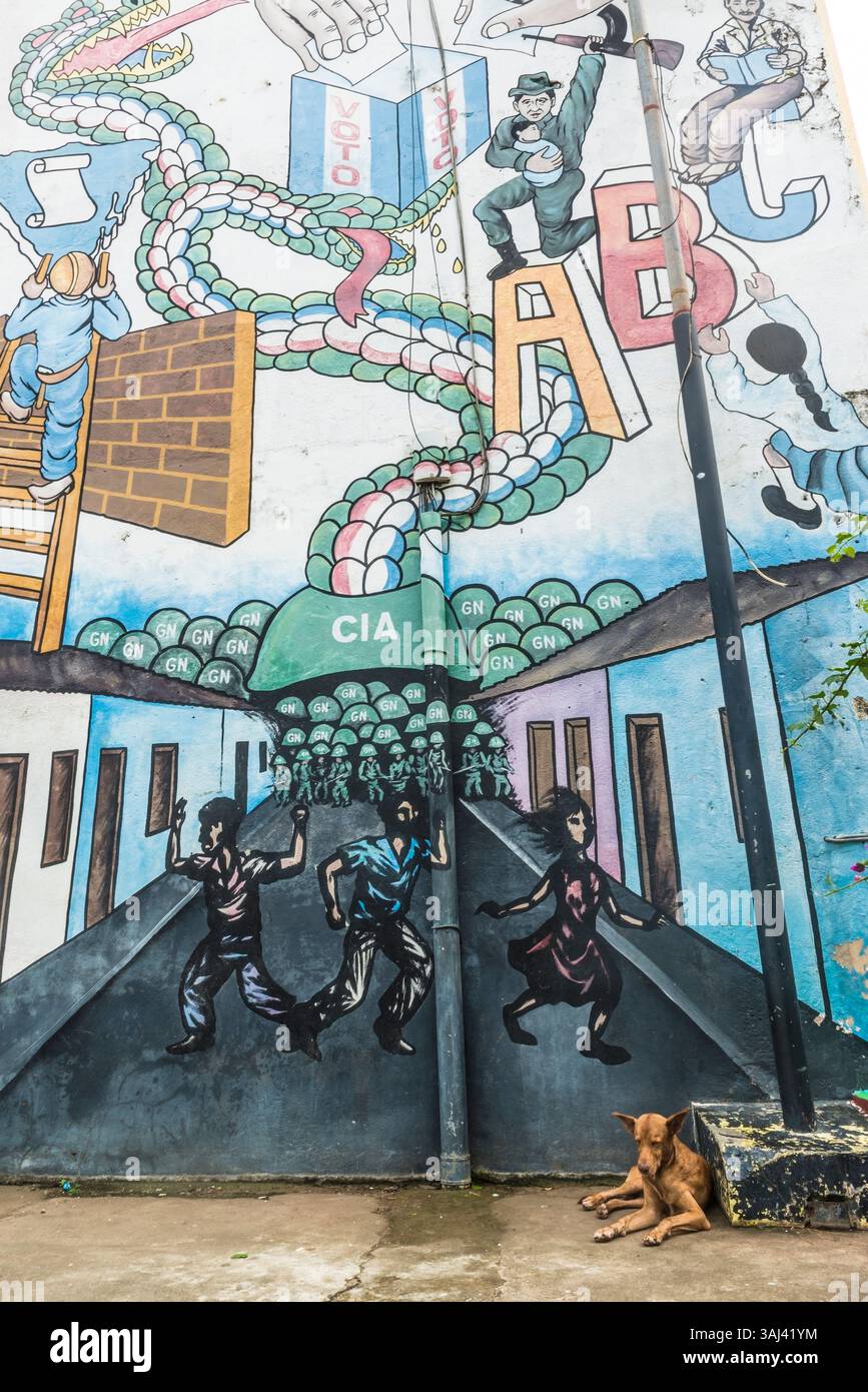 Un cane da strada marrone giace di fronte a un murale politico sull'oppressione del regime di Somoza sostenuto dagli Stati Uniti. Leon, Nicaragua. Foto Stock