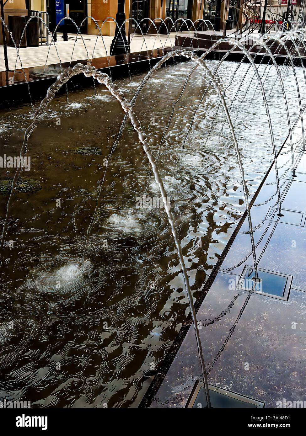 Corsi d'acqua ad arco spruzzano da una fontana moderna in una piazza della città, che mostra simmetria, movimento e riflessi in un ambiente urbano. Foto Stock