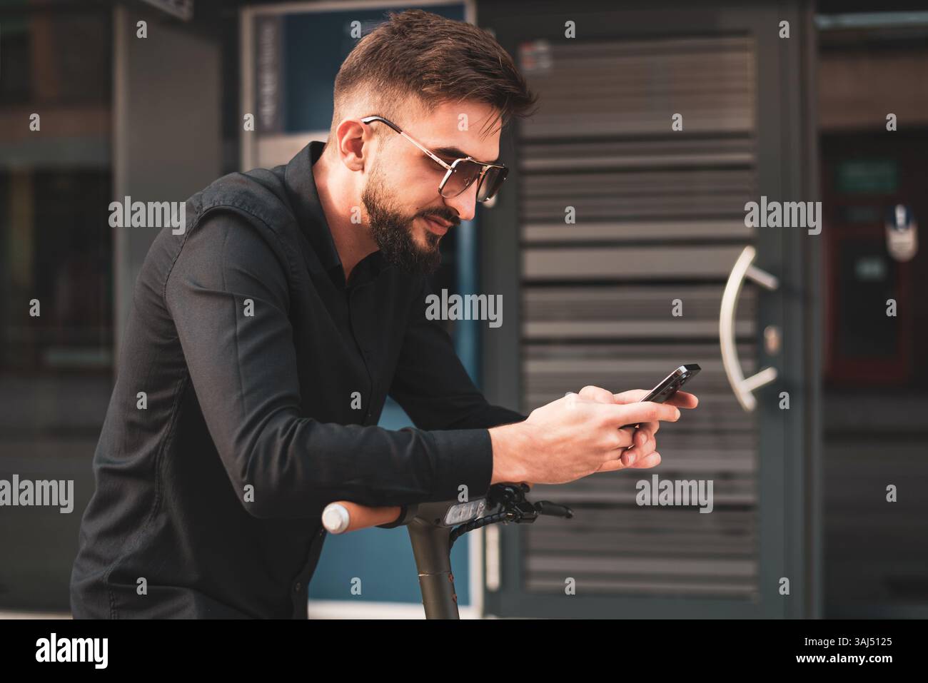 Un giovane uomo urbano elegante con scooter elettrico in piedi di fronte al moderno edificio aziendale sulla strada della città e utilizzando un telefono. Foto Stock