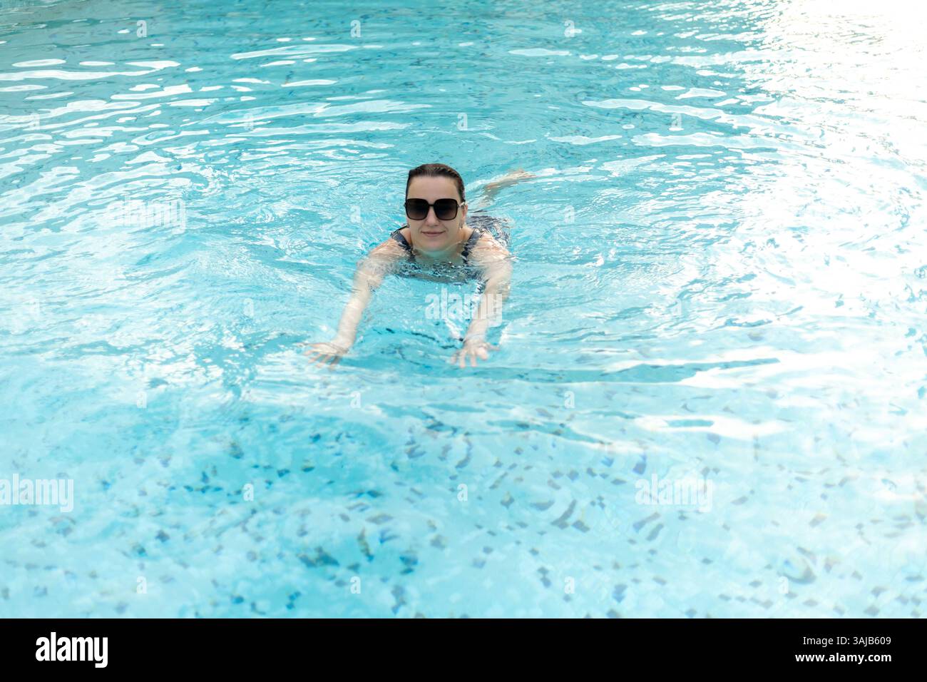 Una giovane donna ama nuotare in una piscina azzurra sotto il sole. Indossa eleganti occhiali da sole e sembra rilassata mentre si muove attraverso l'acqua. Foto Stock