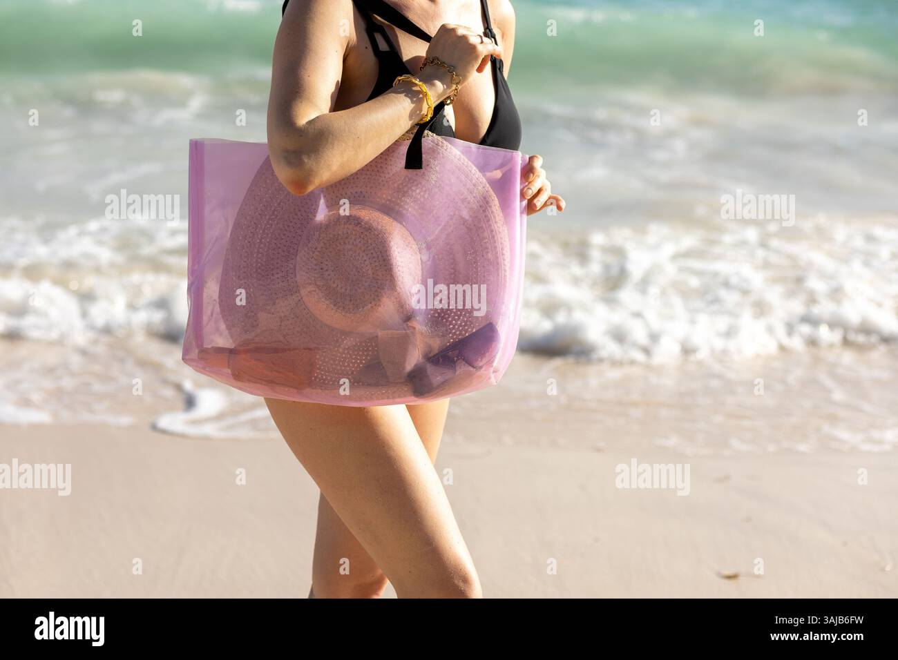 Una donna passeggia sulla morbida spiaggia di sabbia, tenendo in mano una borsa alla moda. Indossa un costume da bagno nero, godendosi il sole luminoso e le dolci onde della b Foto Stock
