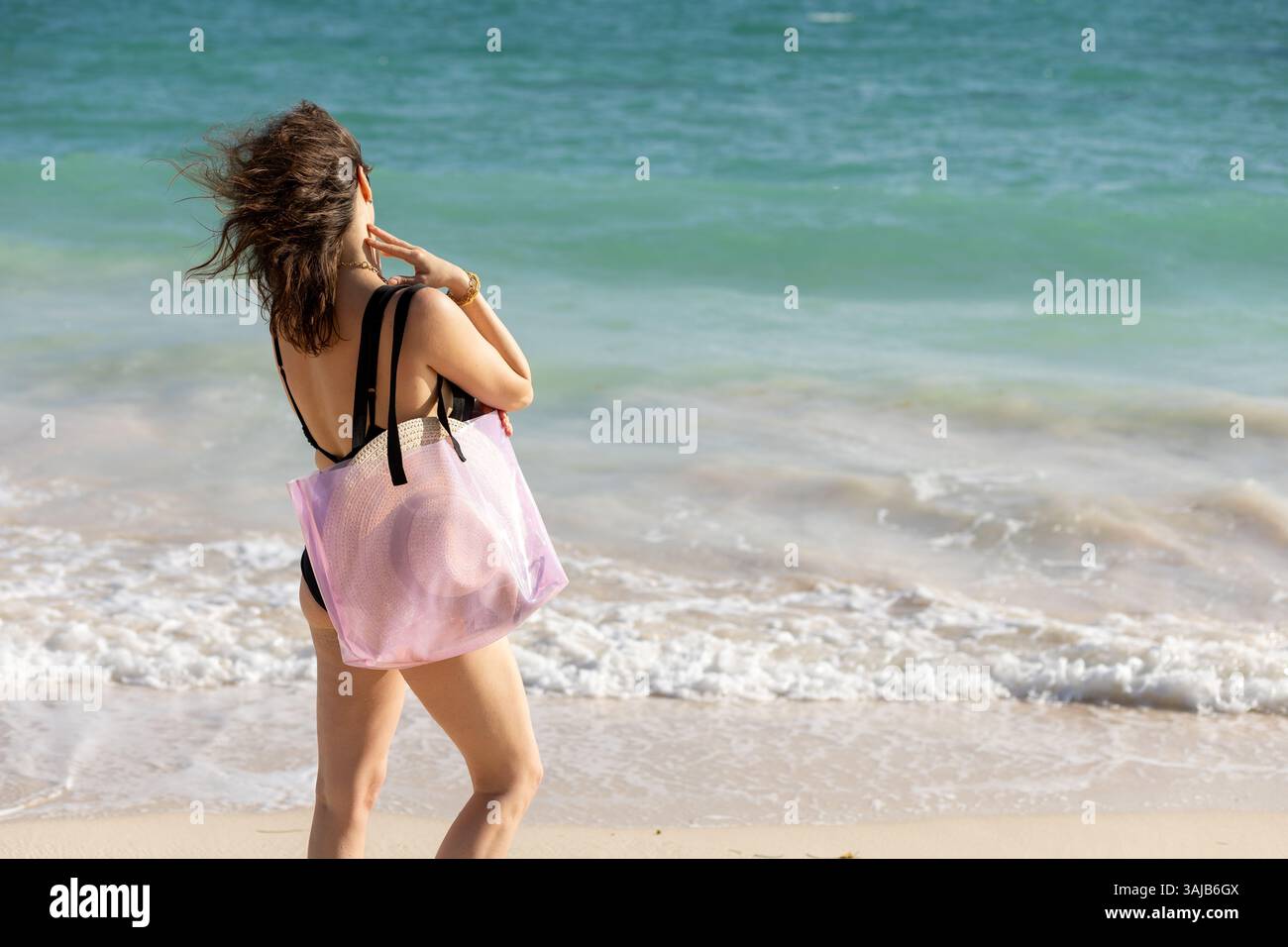 Una donna con i capelli che scorrono passeggia sulla spiaggia, indossa un costume da bagno e porta con sé una borsa tote colorata. Ammira le dolci onde dell'oceano su un caldo sole Foto Stock