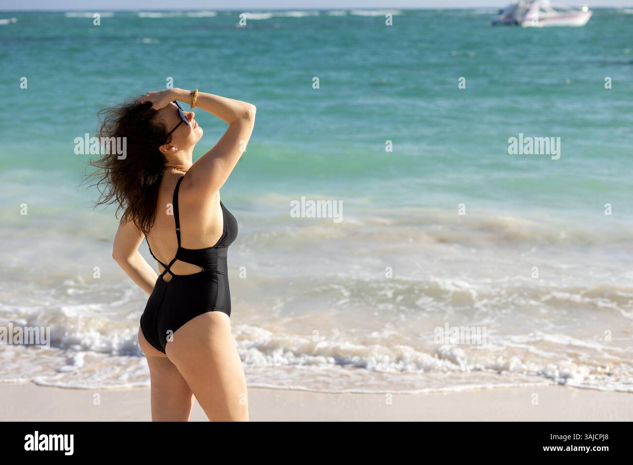 Una donna sta in piedi sulla riva, i suoi capelli sono agitati dalla brezza marina mentre guarda le onde. Indossa un elegante costume da bagno nero, godendosi una piacevole giornata Foto Stock