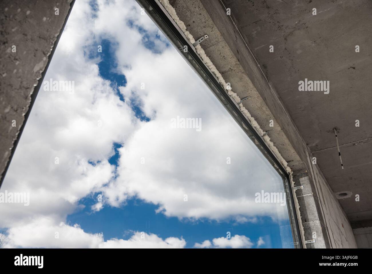 Una grande finestra incornicia una vista del cielo piena di soffici nuvole. La scena cattura il contrasto tra il cemento grezzo dell'edificio infinito Foto Stock