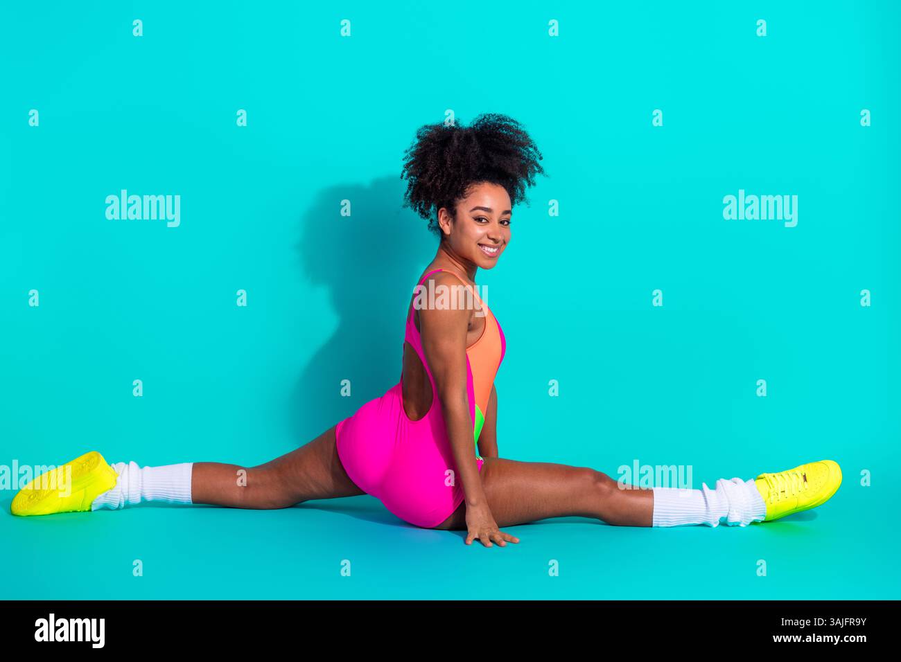Giovane donna atletica che sorride con sicurezza indossando un vivace abbigliamento fitness mentre si mette in posa su uno sfondo turchese brillante Foto Stock