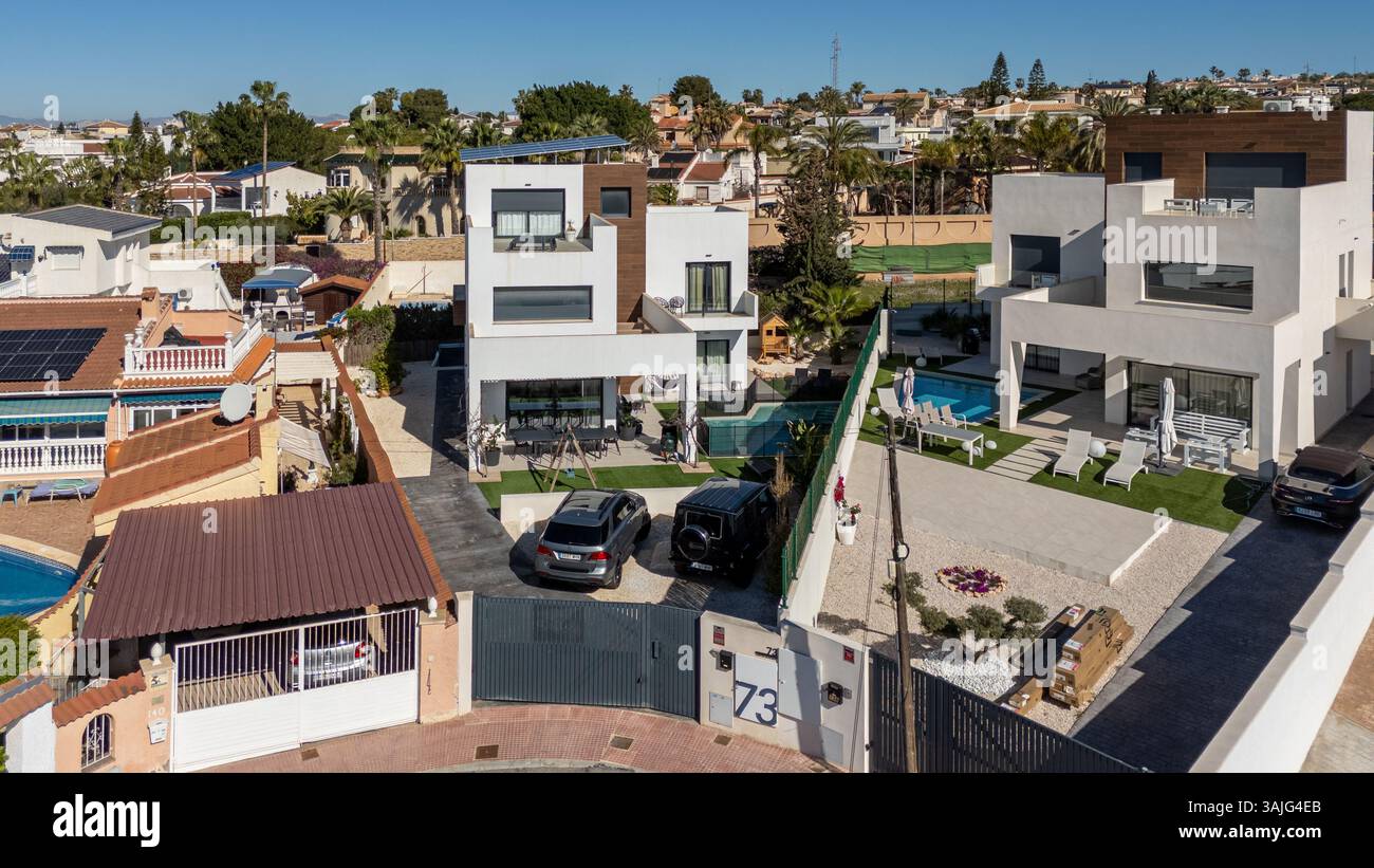 Un'immagine aerea rivolta in avanti che mostra due ville contemporanee con cancelli, vialetti e auto parcheggiate, situate in un soleggiato quartiere residenziale spagnolo Foto Stock