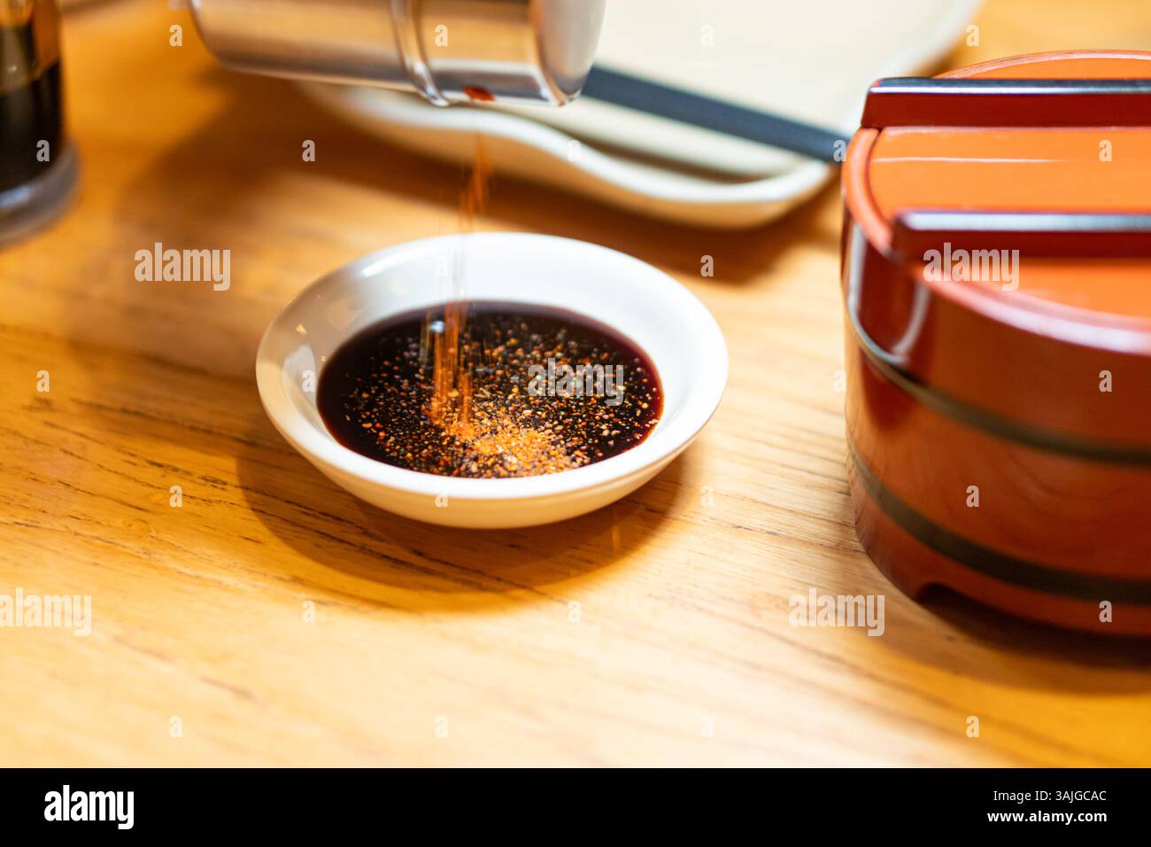 Un piccolo piatto bianco ripieno di salsa di soia scura, con una spolverata di peperoncino rosso. Sullo sfondo, ci sono piatti tradizionali giapponesi Foto Stock