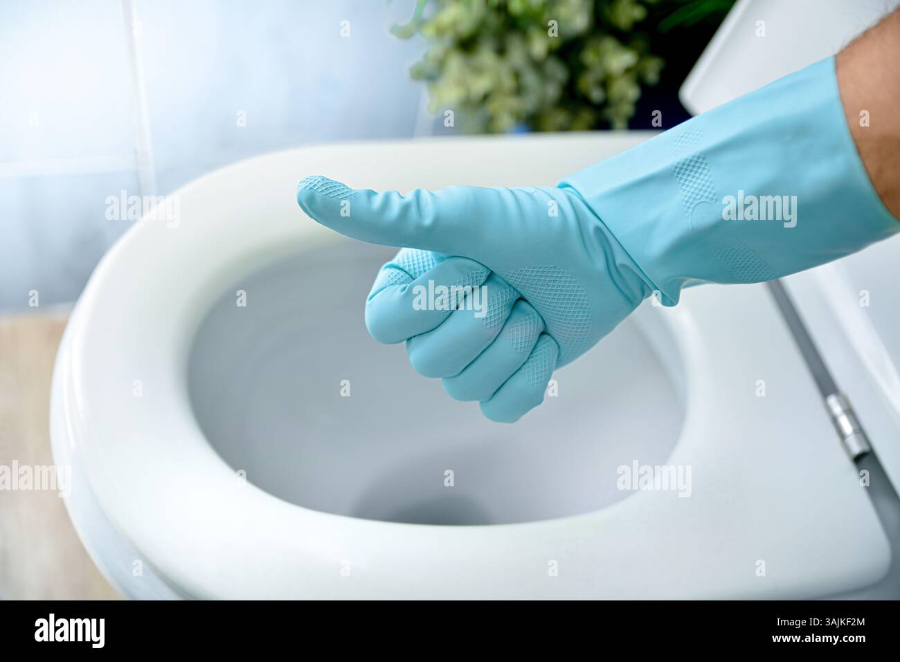 Mano con guanto di gomma blu scrubbing con gesti ok e sfondo con ciotola igienica appena disinfettata con coperchio aperto in un bagno blu. Via sopraelevata Foto Stock
