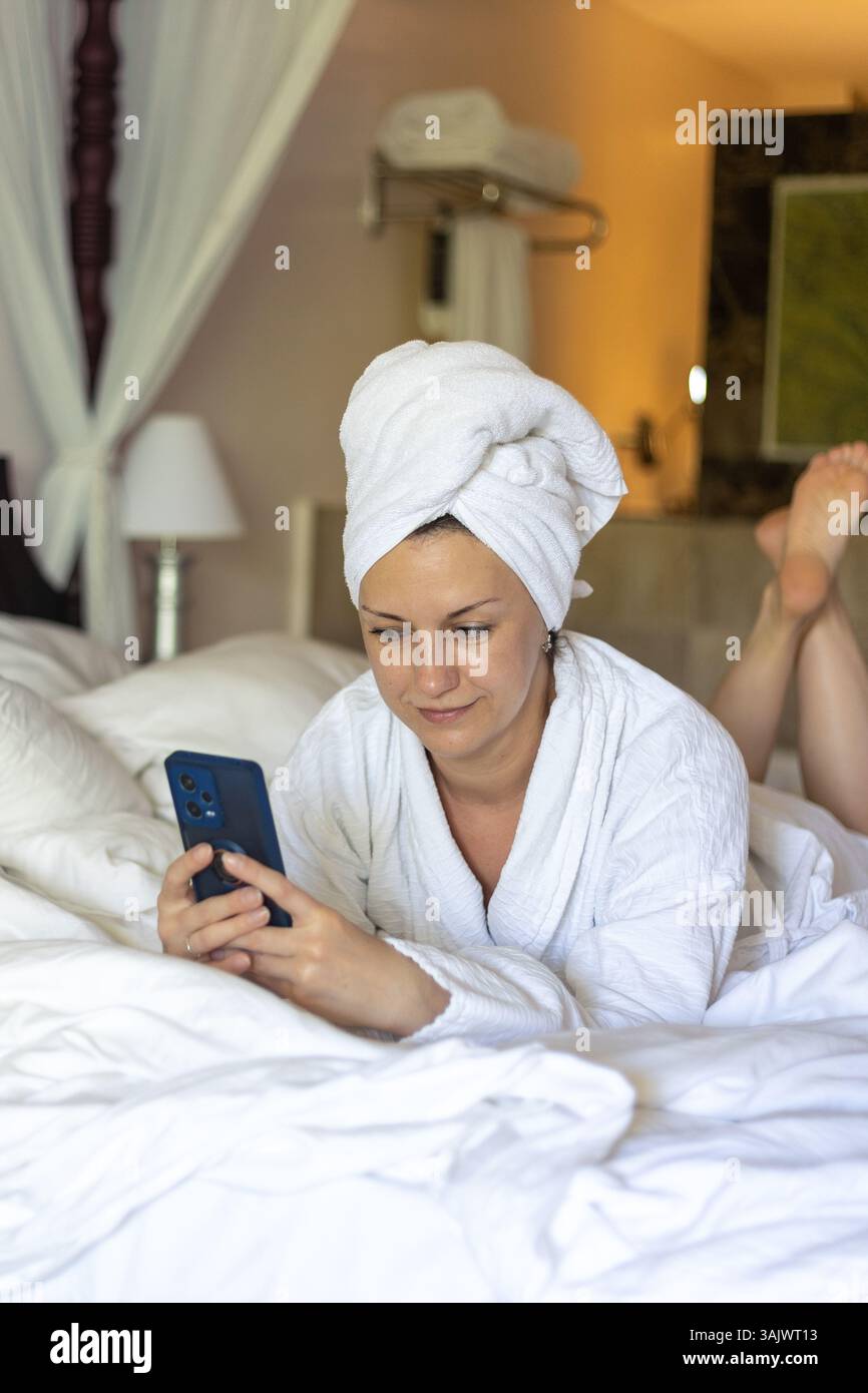 Una donna avvolta in un accappatoio bianco si stende comodamente su un letto, concentrata sul suo smartphone in una camera d'hotel ben illuminata. L'atmosfera è serena e rilassante Foto Stock