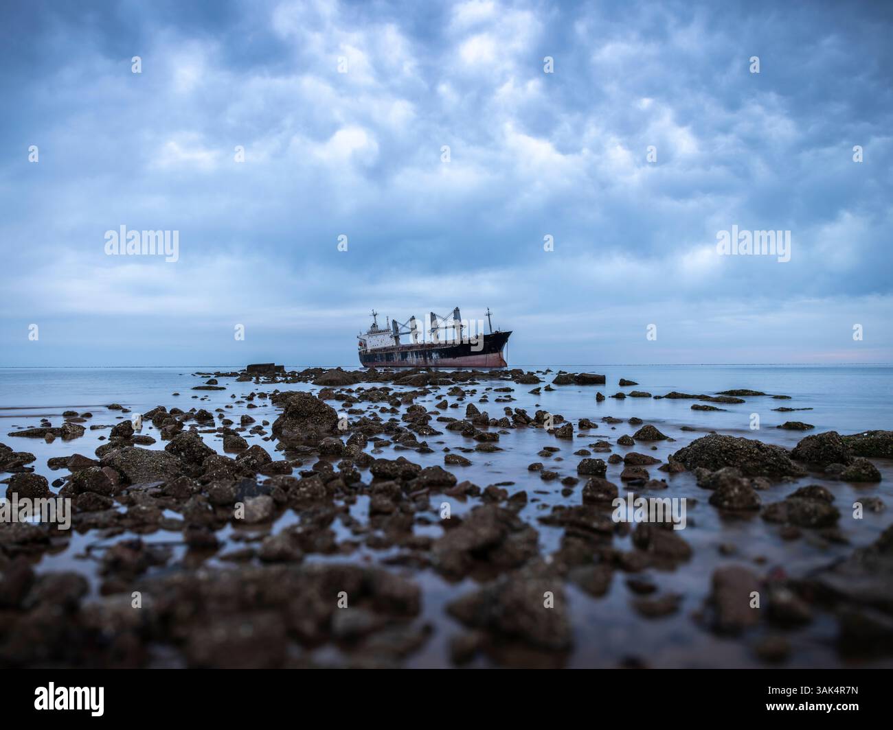 Una nave arrugginita riposa abbandonata sulla costa rocciosa sotto cieli ripidi, scafo intemprato che contrasta rocce frastagliate in tonalità fredde Foto Stock