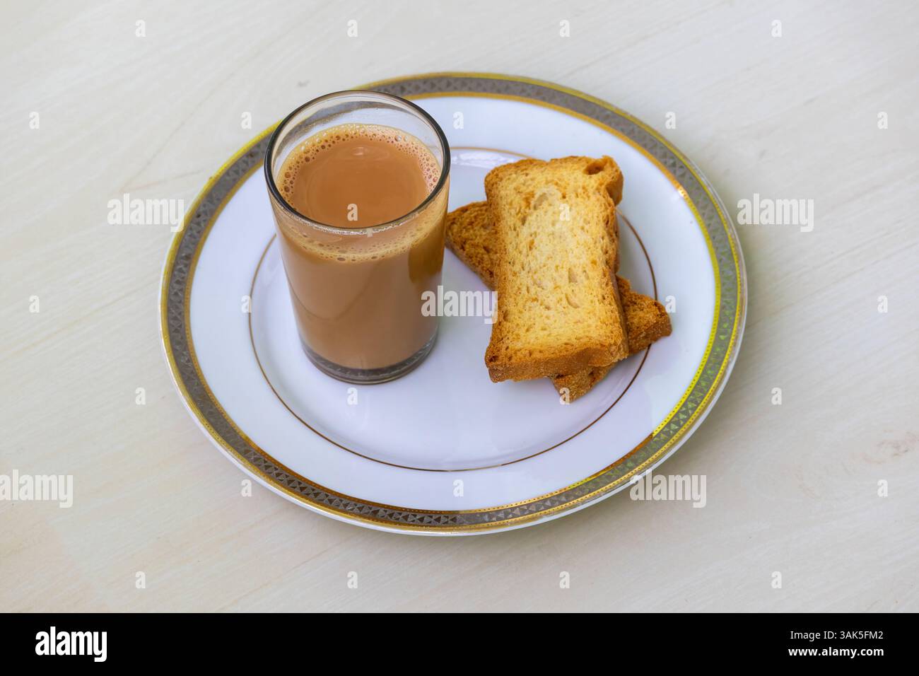 Tè al latte caldo (dudh cha) in una tazza di bicchiere e due pezzi di pane tostato croccante e gustoso (o biscotti al croccante) su un piatto di ceramica su un tavolo di legno chiaro. Foto Stock