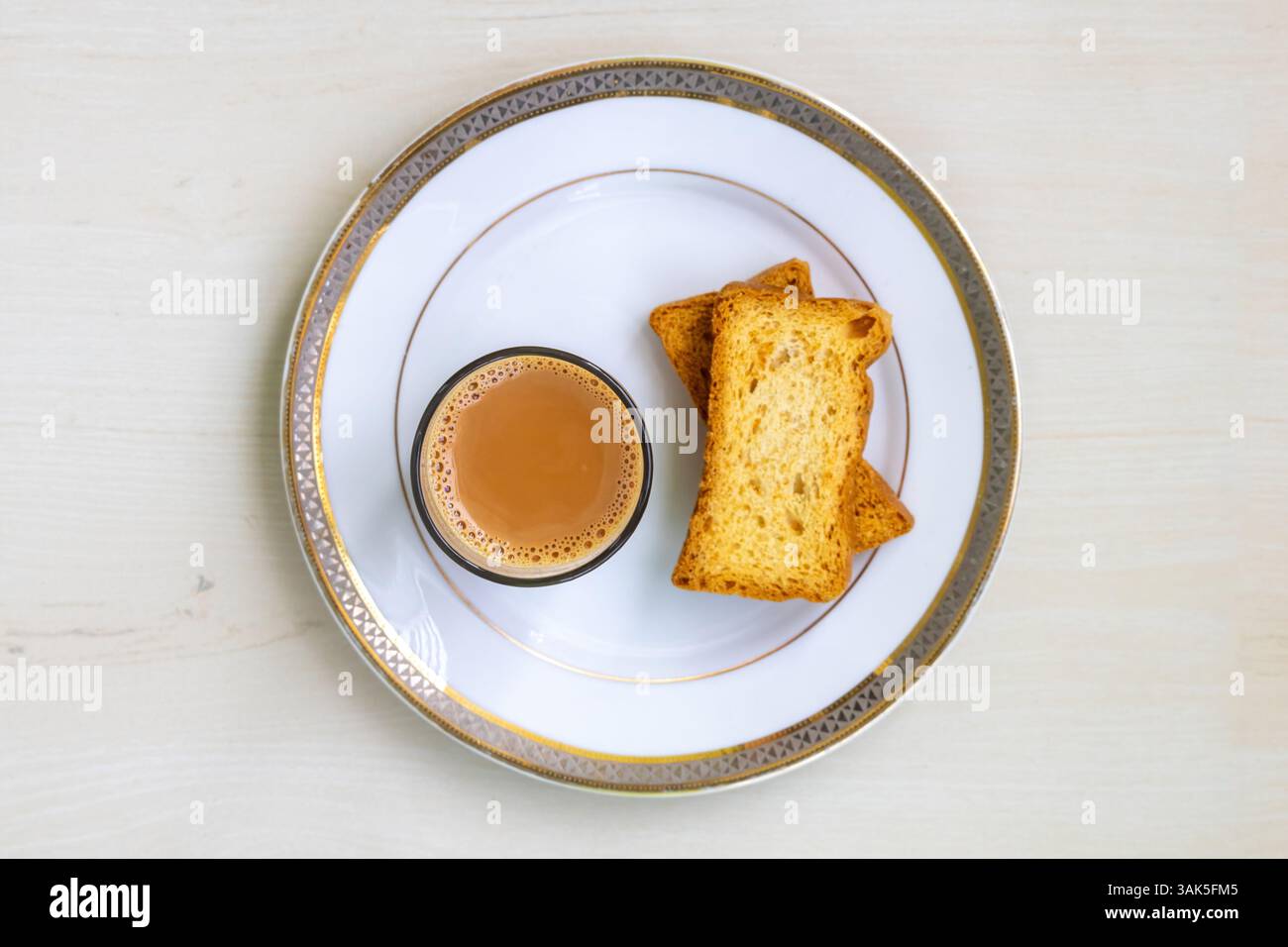 Una tazza di tè al latte caldo (dudh cha) e due pezzi di pane tostato croccante e gustoso (o biscotti al croccante) poggiano su un piatto di ceramica su un tavolo di legno chiaro. V superiore Foto Stock