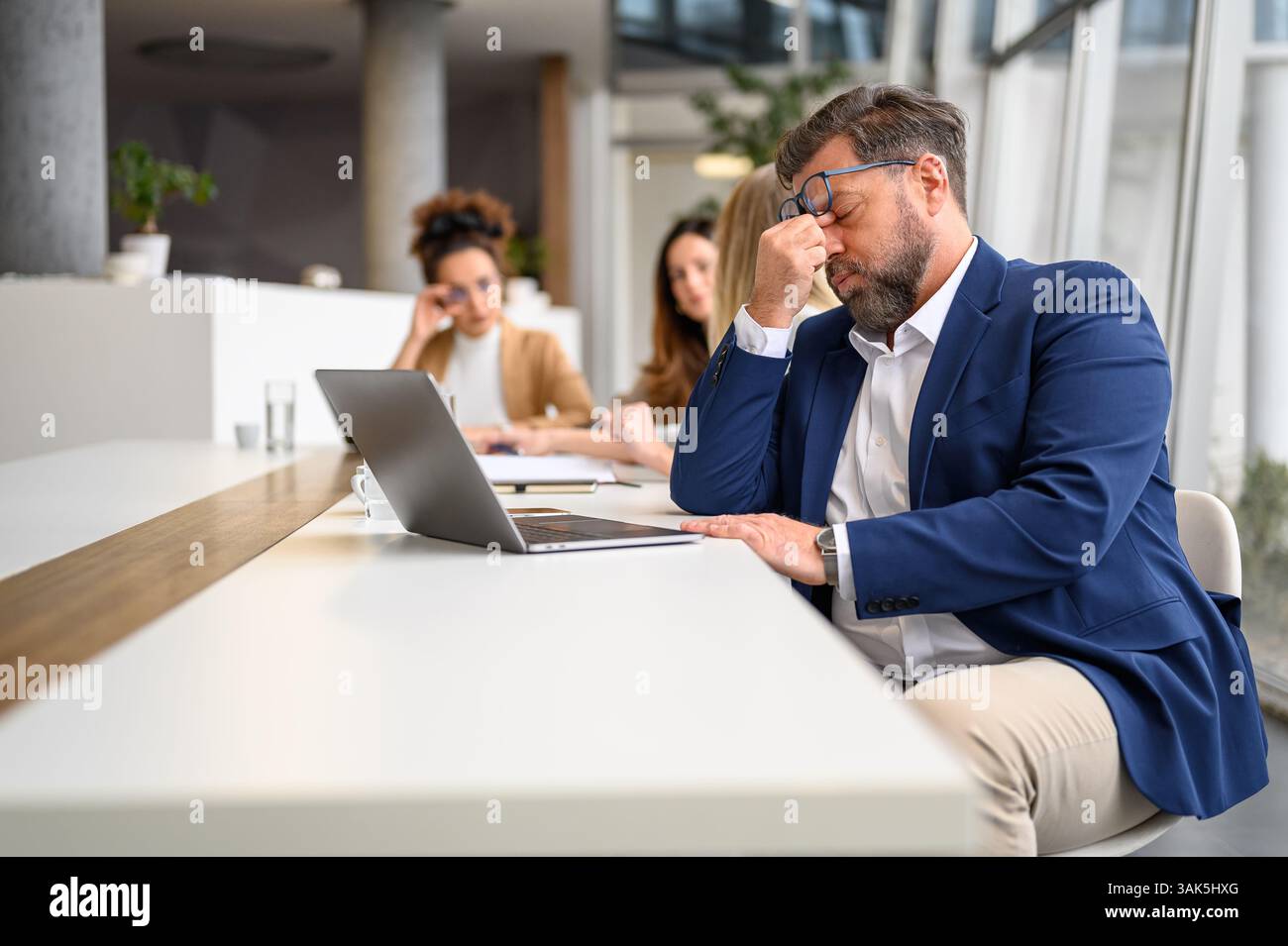 Un uomo d'affari stressato si strofina gli occhi mentre lavora su un notebook con colleghi di sesso femminile sullo sfondo Foto Stock