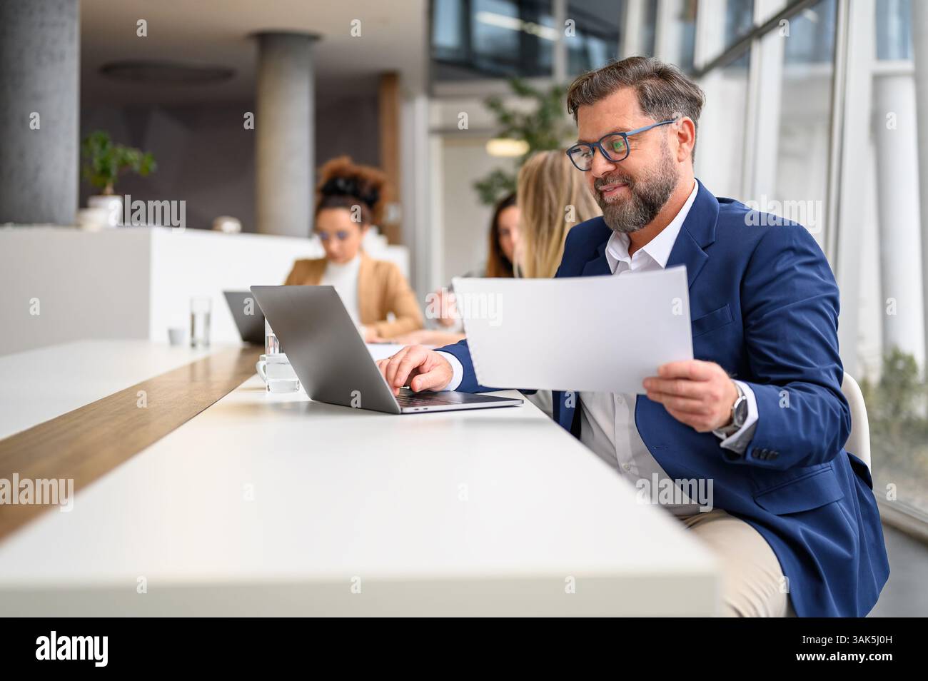 Uomo d'affari che analizza i documenti mentre lavora su un computer portatile in un ufficio moderno con colleghi di sesso femminile in background Foto Stock