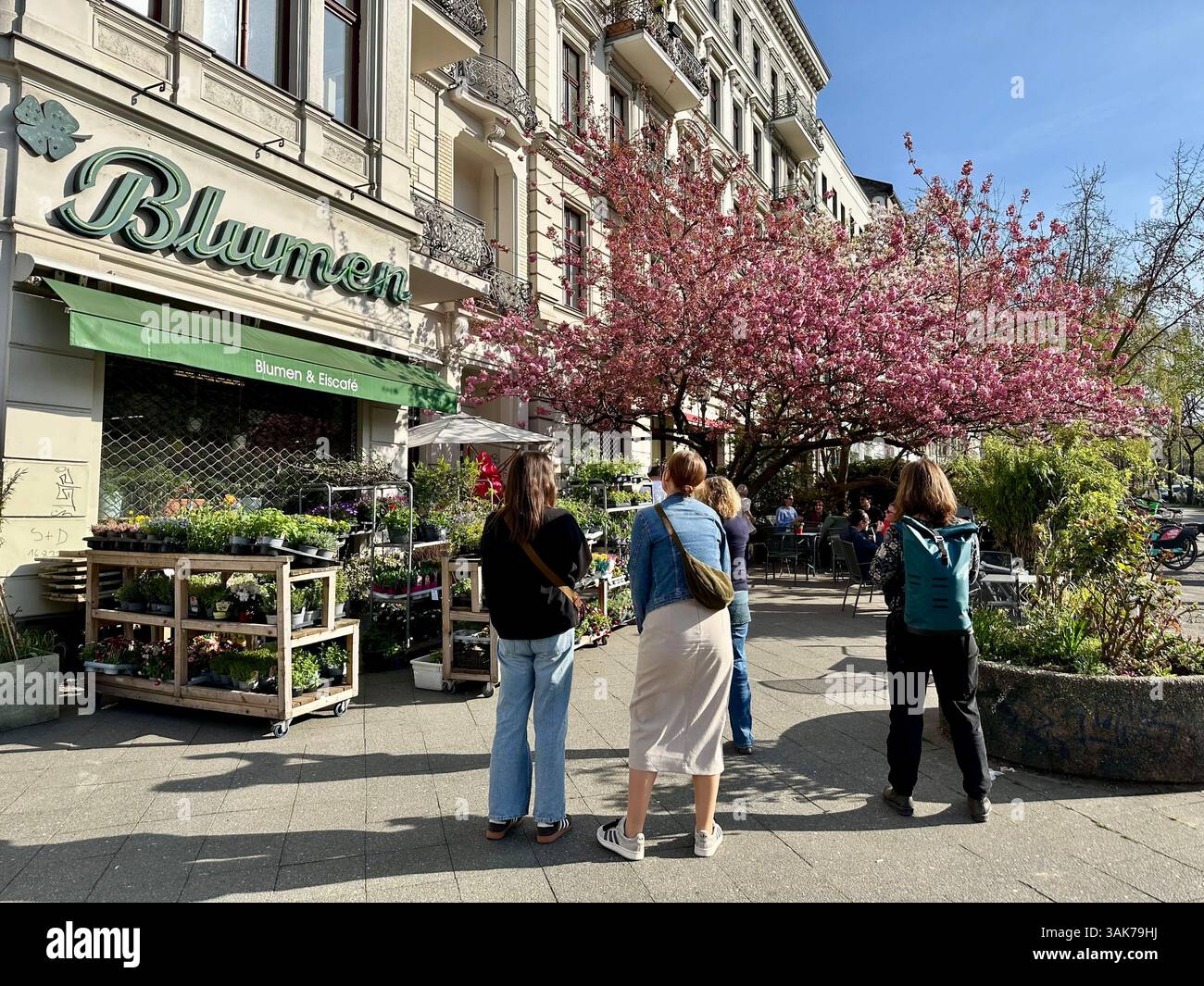 Persone in piedi di fronte a un negozio di fiori che ammirano la fioritura di un ciliegio in primavera, Kreuzberg, Berlino - Immagine stock catturata con smartphone