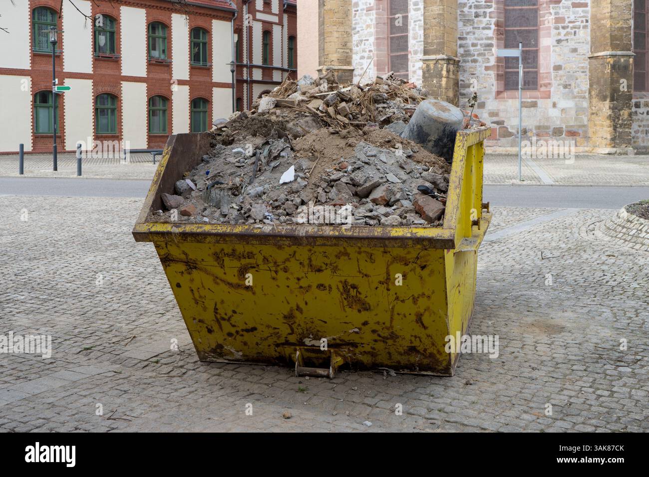 Un contenitore di metallo giallo pieno di detriti da costruzione si trova in una piazza di fronte agli edifici storici Foto Stock