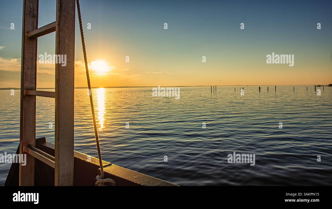 Una scala di legno e una corda di ormeggio incorniciano una tranquilla scena del tramonto sul lago con una luce dorata soffusa che si riflette sulla tranquilla acqua ondulata. Foto Stock