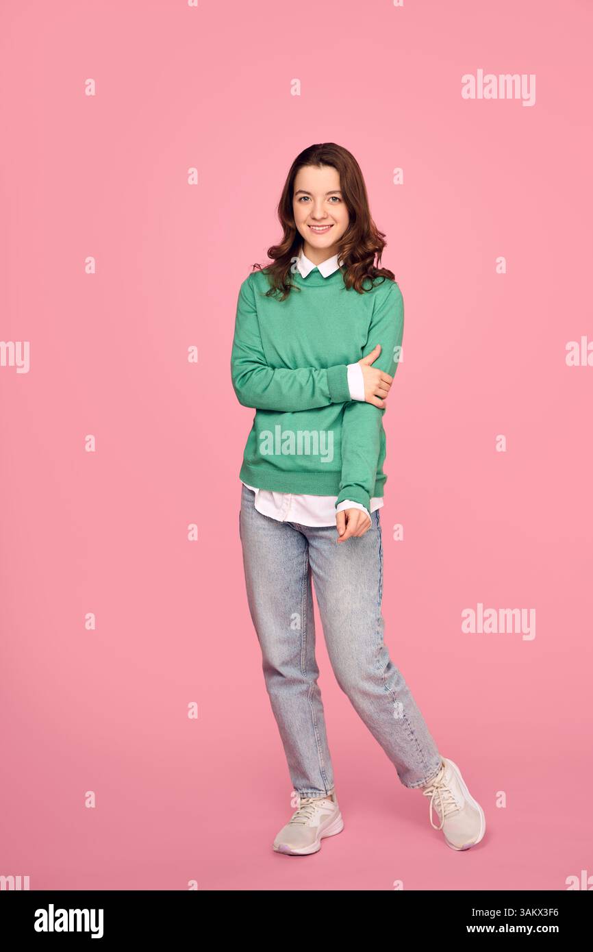 Foto a tutta lunghezza di una giovane donna felice che indossa jeans e un maglione verde isolato su sfondo rosa. Foto Stock