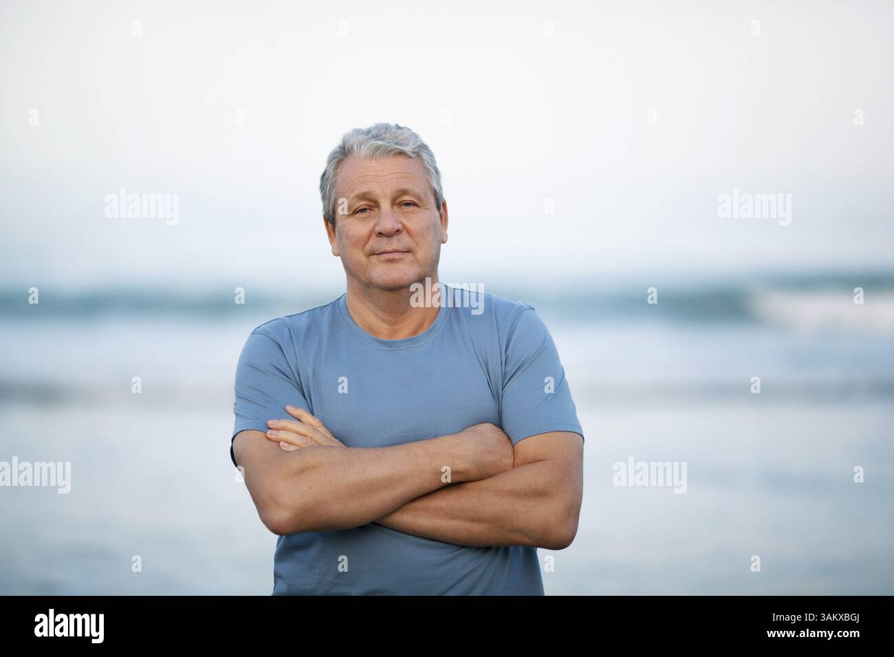 Immagine centrale di un uomo anziano in maglietta blu con le braccia incrociate sul petto. Persona che guarda la fotocamera con un sorriso leggero. Guarda contro sfocature Foto Stock