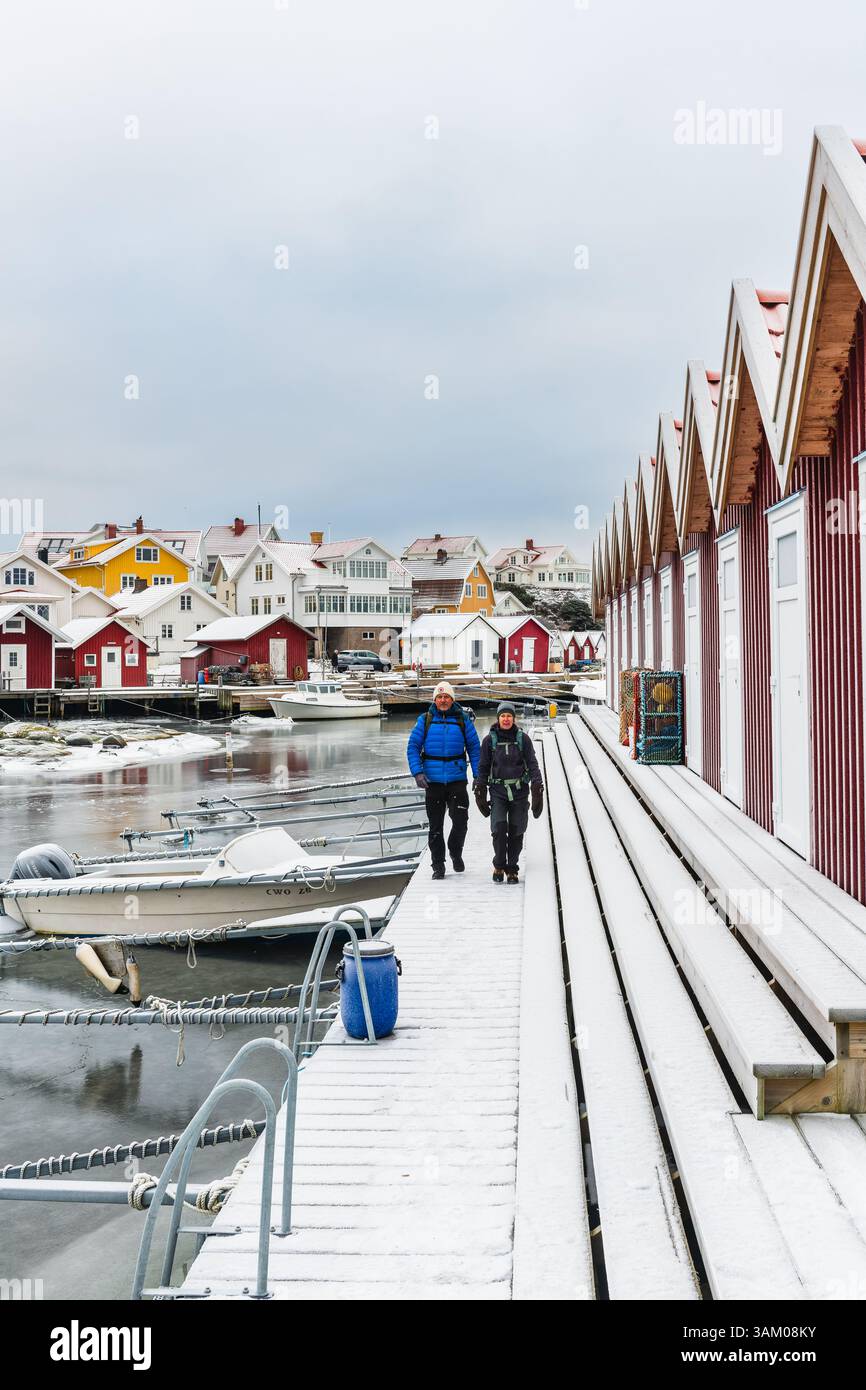 Due escursionisti passeggiano lungo un molo coperto di neve accanto a un pittoresco porto di Gothenburg. Case colorate offrono uno sfondo affascinante contro l'inverno Foto Stock