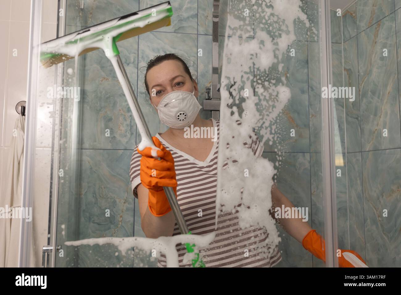 Una donna che indossa una maschera e guanti pulisce accuratamente la cabina doccia con acqua e sapone per garantire igiene e sicurezza Foto Stock
