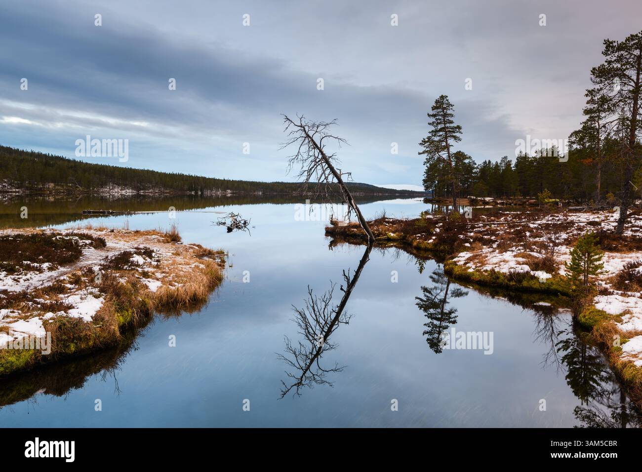 Le acque calme riflettono gli alberi lungo la riva e un paesaggio lontano in Svezia. L'ambientazione invernale include macchie di neve che contribuiscono alla tranquillità Foto Stock