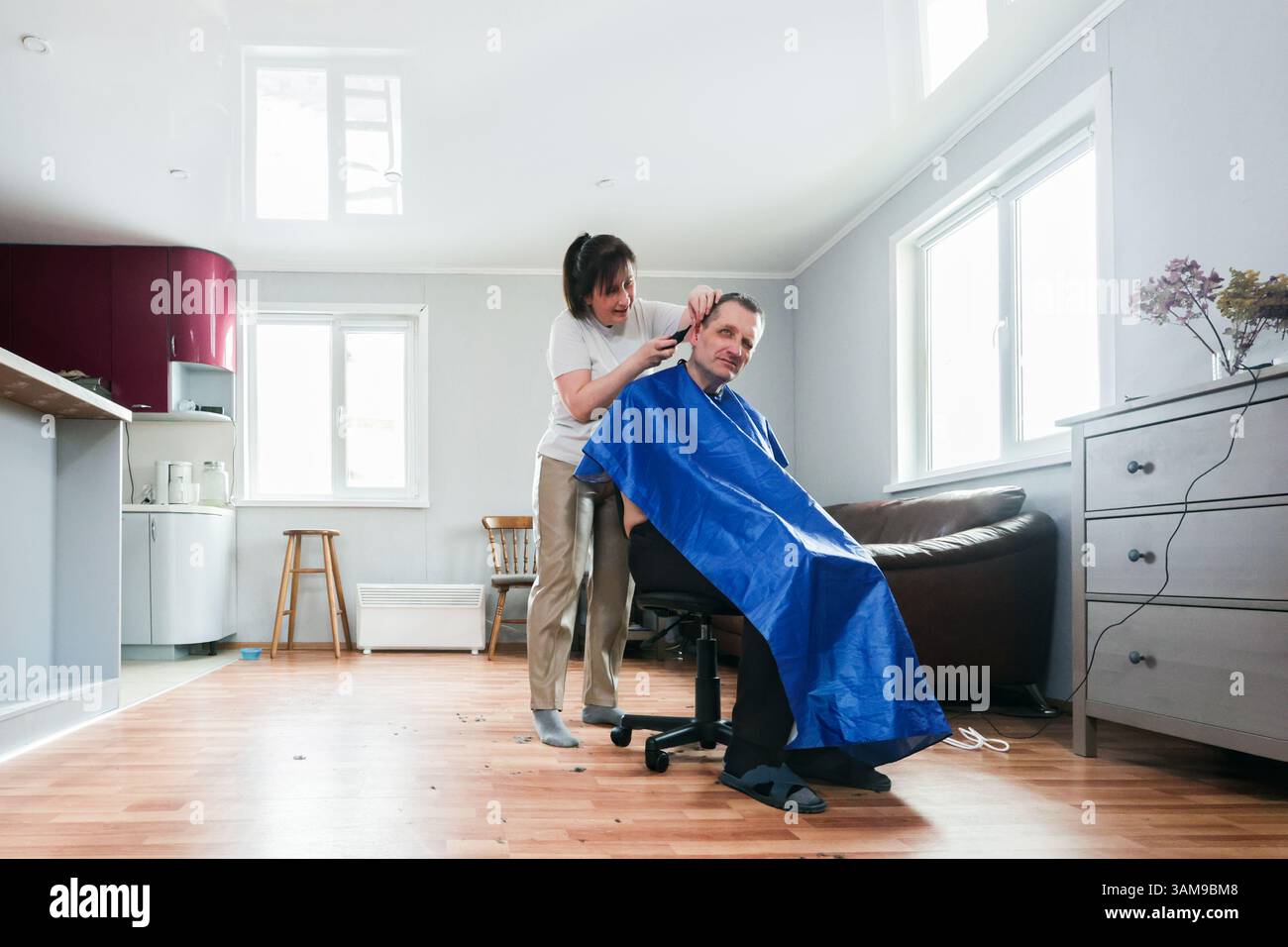 Il coniuge sta completando il taglio di capelli rifilando le orecchie e il naso del partner negli anni '50, in interni, con capelli sparsi su pavimento in legno e luce naturale dalla finestra. Foto Stock