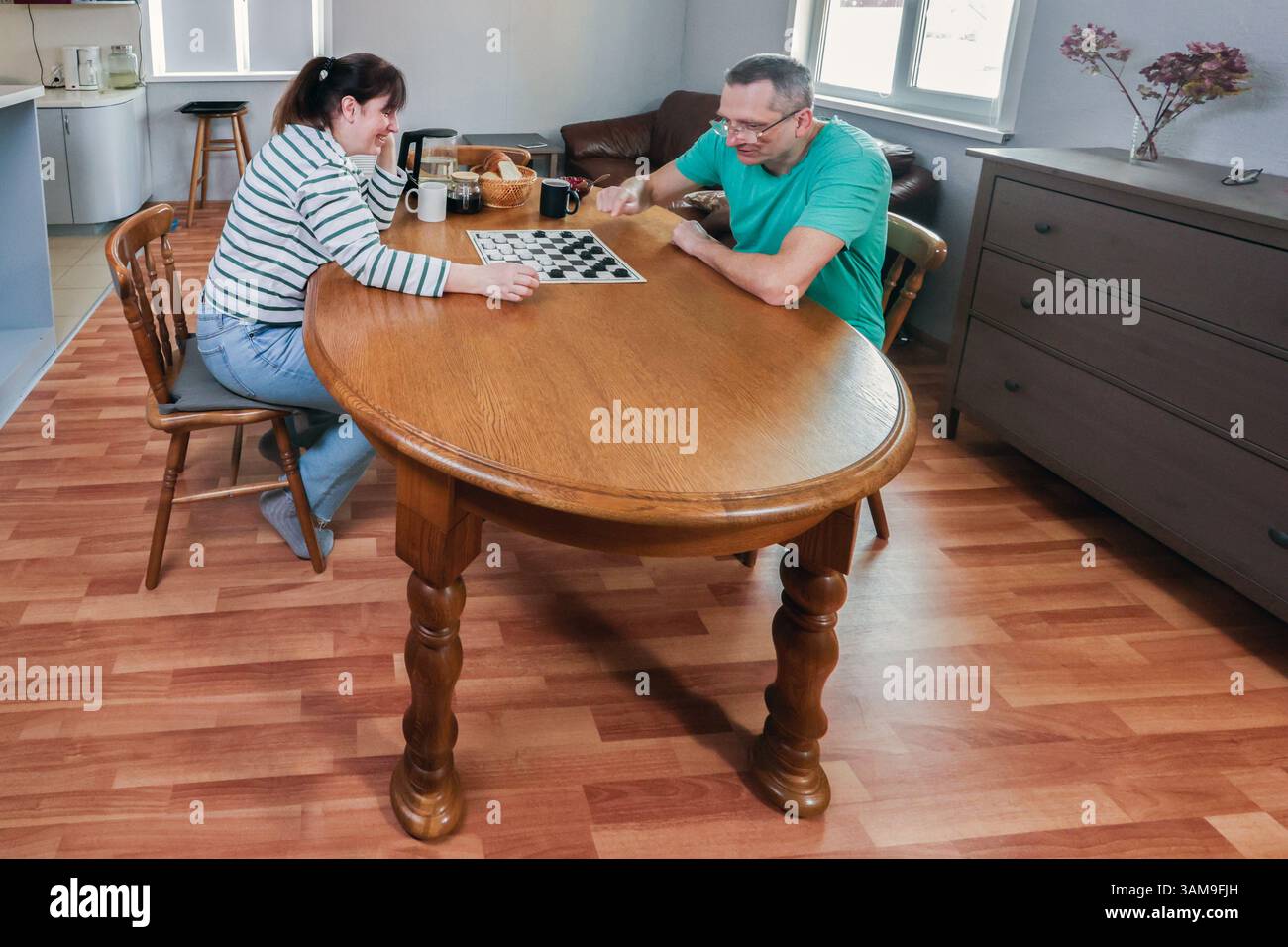 Adulti maturi sono impegnati in una partita di dama a un grande tavolo, circondati da mobili di casa, godendosi momenti tranquilli in un ambiente interno luminoso Foto Stock