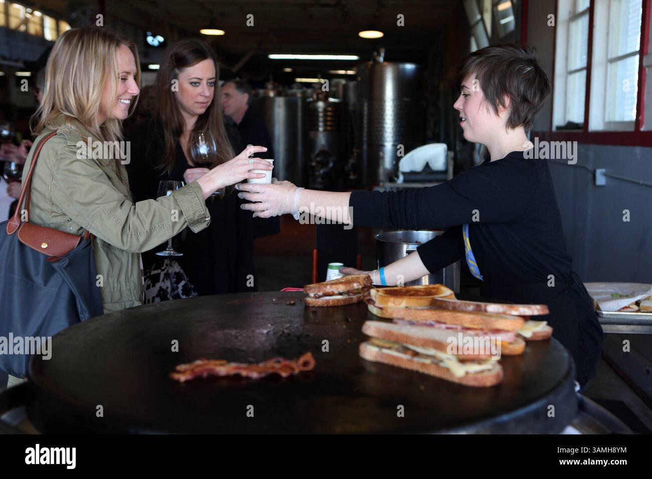 21 dicembre 2013 - San Francisco, CA, USA - dipendente Melanie Rose, Right, of American Grilled Cheese Kitchen, consegna la zuppa di pomodoro affumicata mentre i panini vengono cucinati alla griglia durante l'evento Winter Solstice Wine Tasting and Grilled Cheese presso Treasure Island Wines on Treasure Island a San Francisco il 21 dicembre 2013. Treasure Island Wines condivide il magazzino con altre aziende vinicole; vini di cuore, vini Eristavi, vini Bravium/artigianali e vini recentemente Conduit. (Immagine di credito: © Ray Chavez/MCT/ZUMAPRESS.com) Foto Stock
