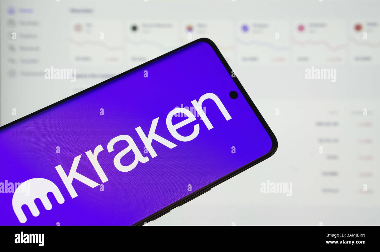 il logo kraken viene visualizzato sullo schermo di uno smartphone, con una pagina web del grafico delle azioni visibile sullo schermo di un computer sullo sfondo Foto Stock