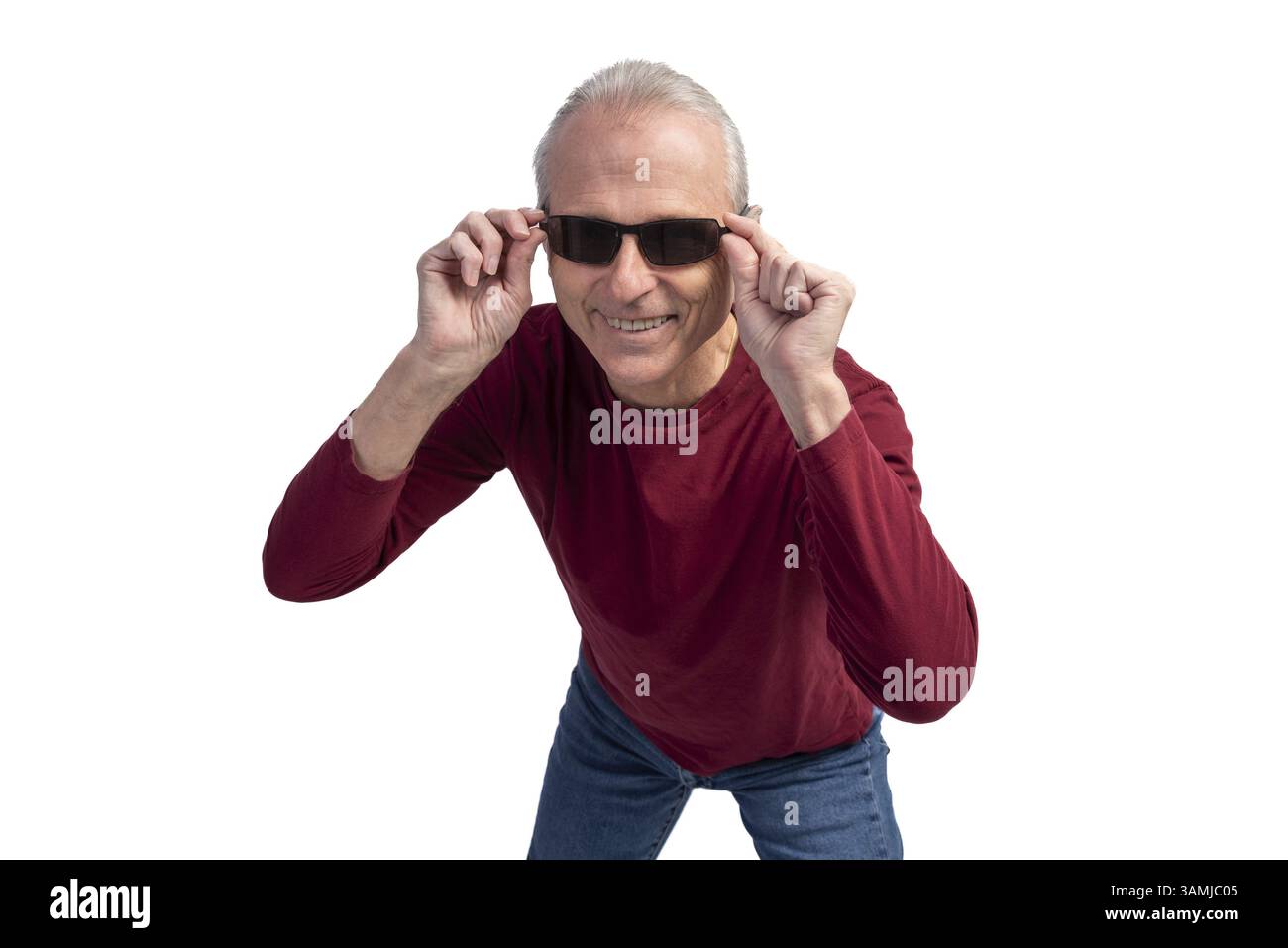Uomo anziano giocoso in abbigliamento casual, occhiali da sole abbassati con un sorriso caldo, che emana gioia e positività su uno sfondo bianco brillante Foto Stock