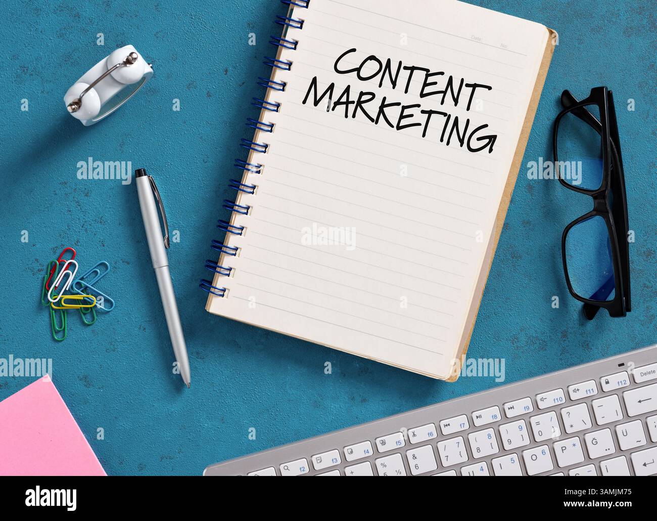 Blocco note con contenuto marketing scritto su di esso, posizionato su una scrivania blu con tastiera, penna, orologio, graffette, occhiali, e appunti, che suggeriscono una s. Foto Stock