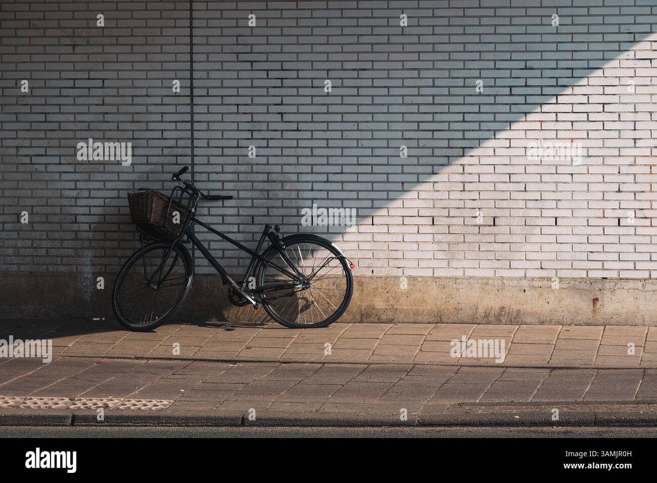 Una bicicletta solitaria con un cesto anteriore si appoggia contro una parete di mattoni bianchi illuminati dal sole su un tranquillo marciapiede urbano. Foto Stock