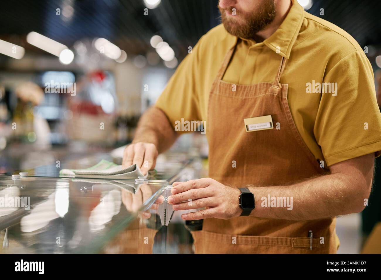 Il dipendente della caffetteria pulisce il piano d'appoggio mentre si concentra sull'attività con l'etichetta del nome visibile. Sfondo che mostra l'ambiente della caffetteria e luci sfocate che creano un'atmosfera movimentata Foto Stock
