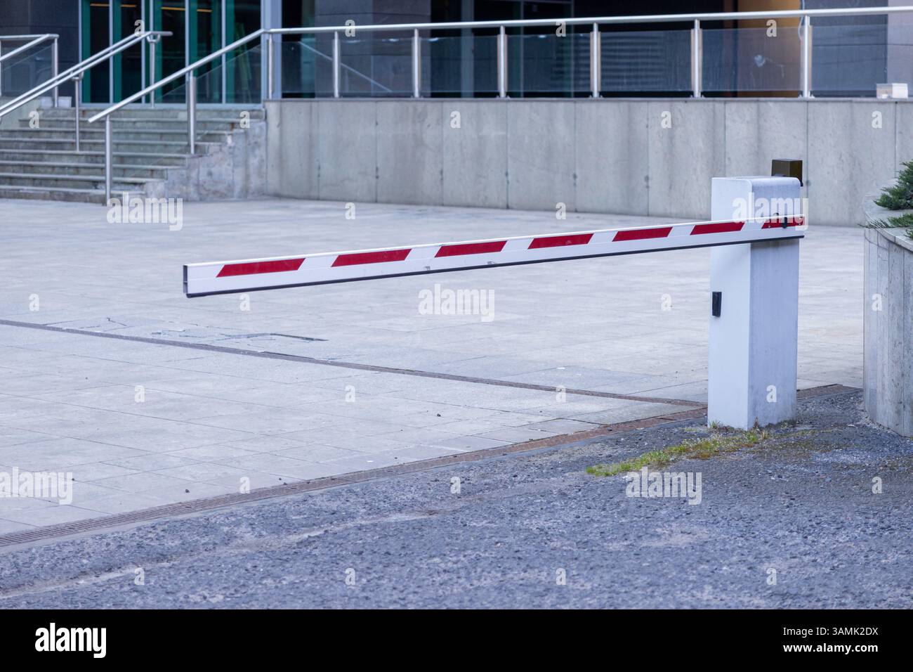 Vista ravvicinata di un cancello di sicurezza a strisce bianche e rosse abbassato attraverso un ingresso pavimentato vicino a un edificio moderno, simboleggiante l'accesso controllato A. Foto Stock