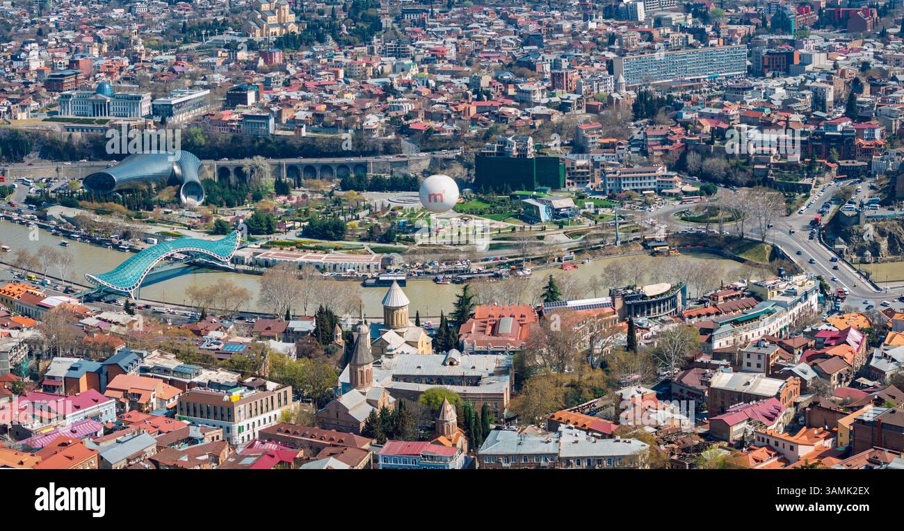 Ampia vista panoramica della città di Tbilisi che si estende lungo il fiume Kura, con ponti, quartieri storici e strutture collinari. Foto Stock